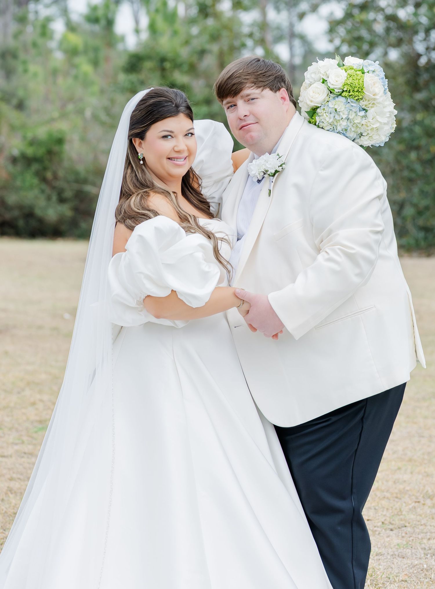 Smiling bride and groom pose together outdoors, bride holding white and blue hydrangea bouquet.