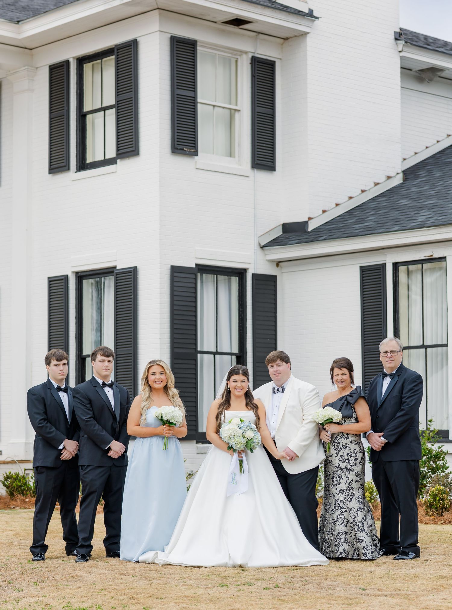 Wedding party poses in front of white Southern mansion with black shutters, bride in white gown holding bouquet.