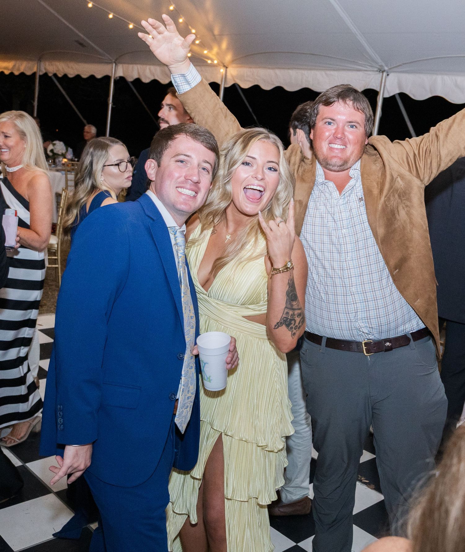Three excited wedding guests posing and celebrating on checkered dance floor at outdoor reception.