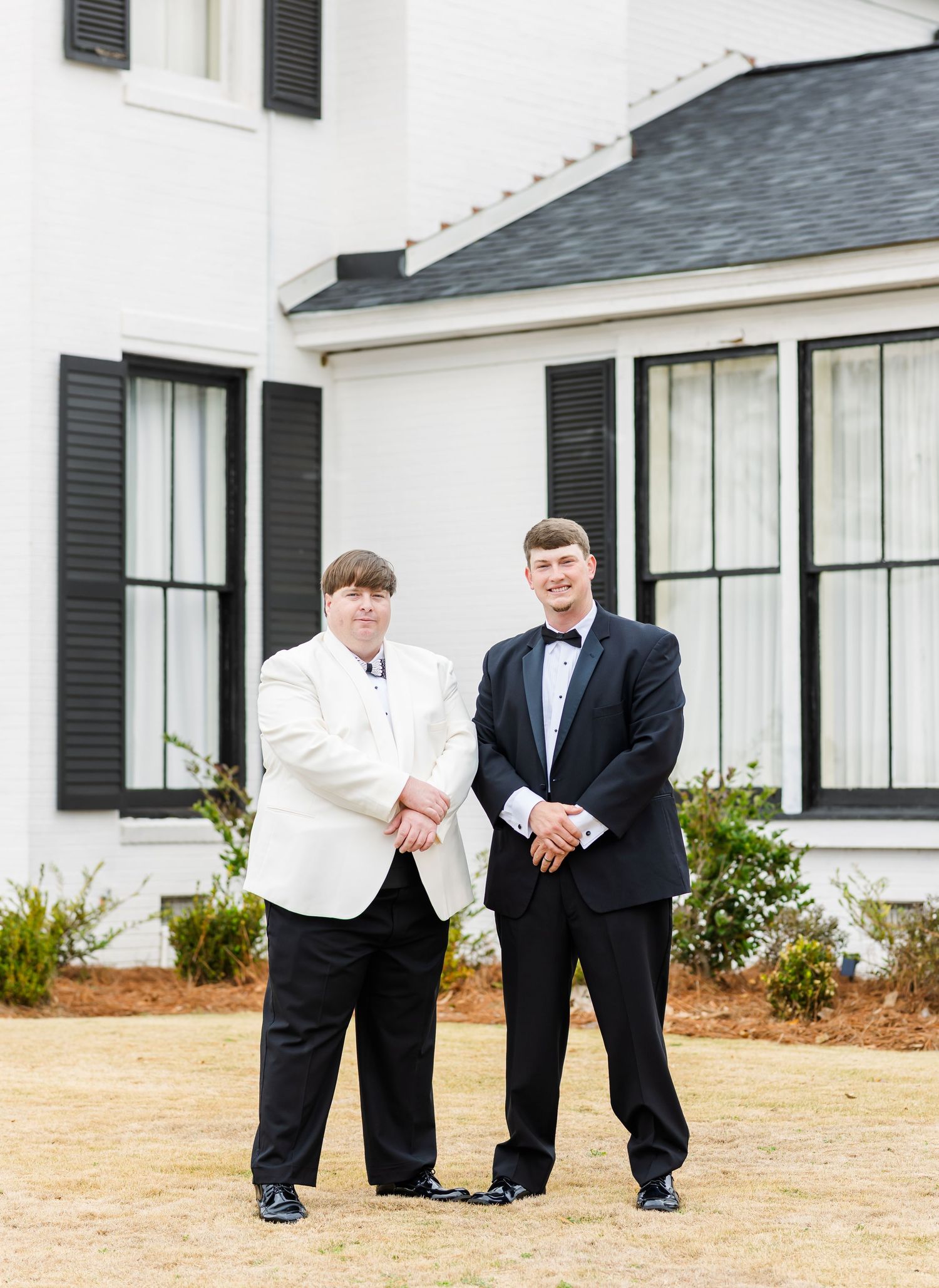Groom in white tuxedo jacket and groomsman in black tuxedo shake hands outside white wedding venue.