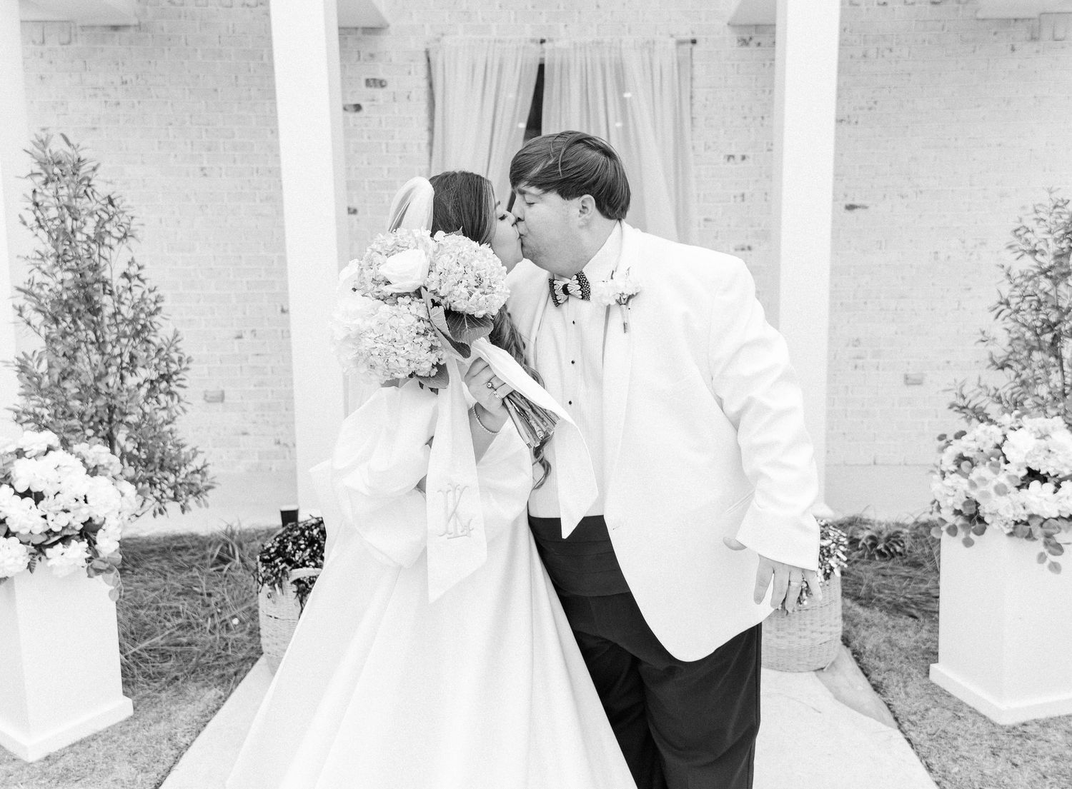 Black and white photo of bride and groom sharing a kiss at outdoor wedding ceremony altar.