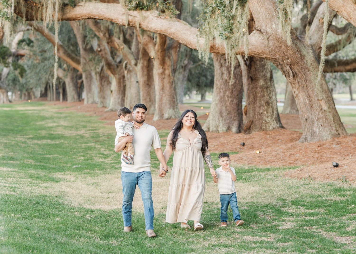 A family walks together holding hands along a tree-lined path in a park on a sunny day.