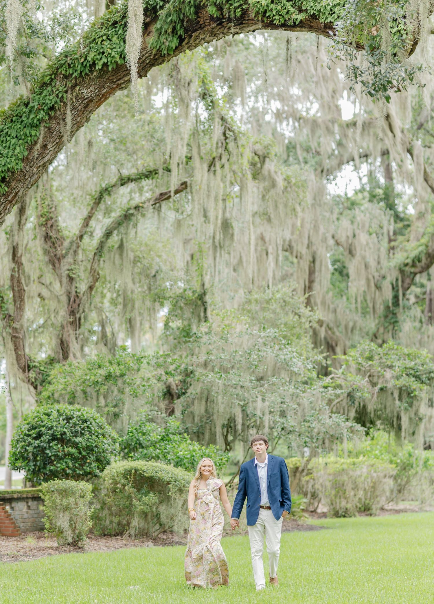 Romantic couple strolling under Spanish moss-draped trees in a garden setting.