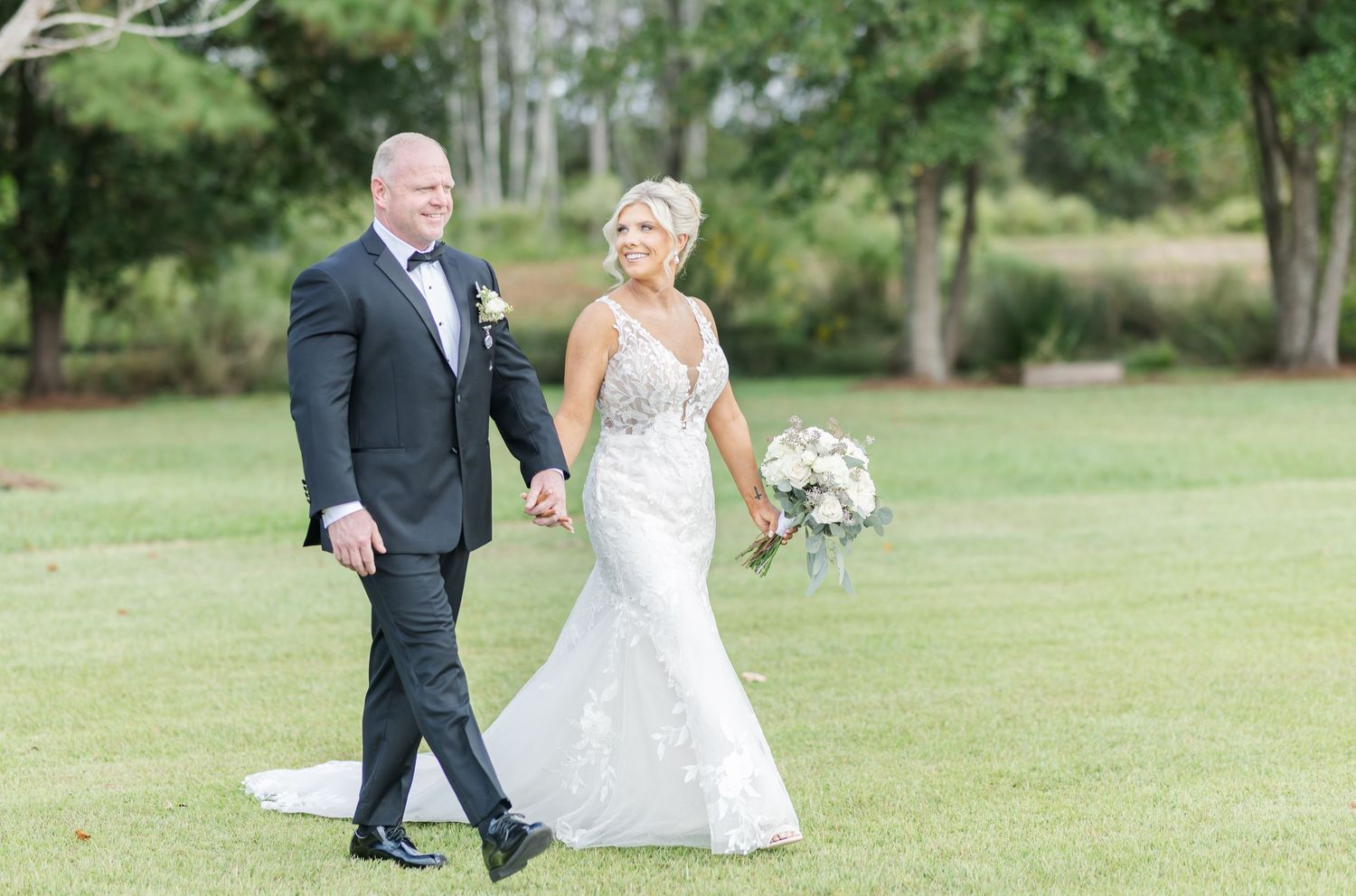A couple walks hand in hand across a lush green lawn during their outdoor wedding celebration.