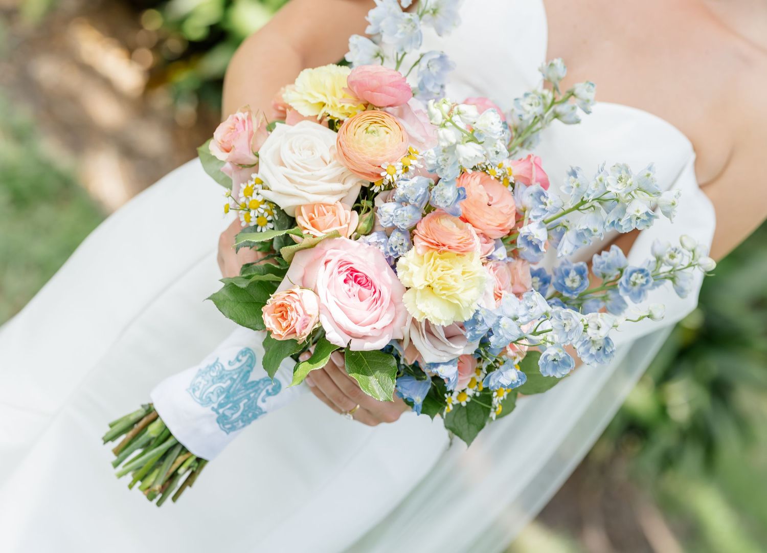A pastel bouquet of peach and pink roses with blue accents rests against a white fabric.