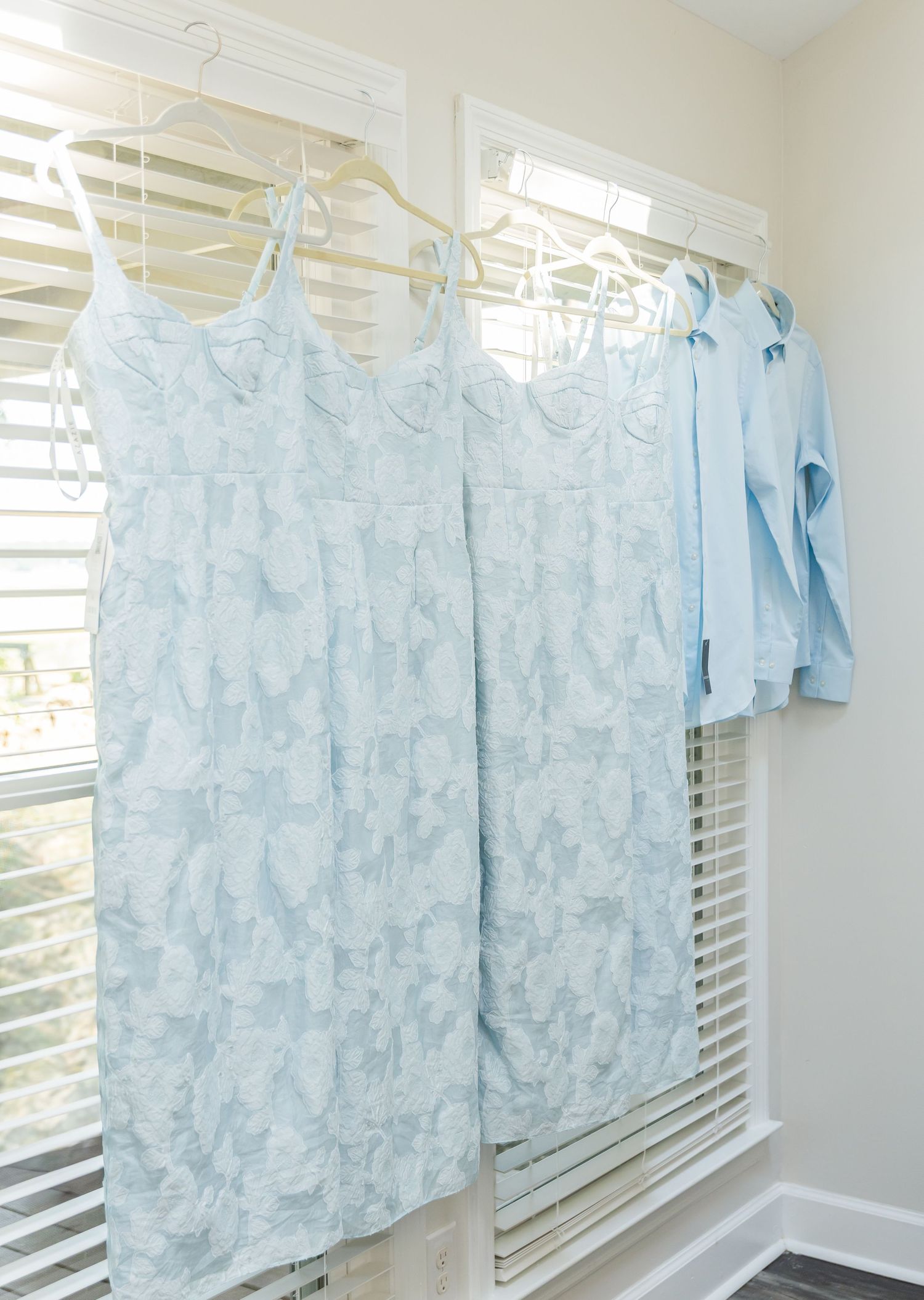 Light blue floral Hawaiian shirts hanging on white hangers against a white wall with venetian blinds in the background.