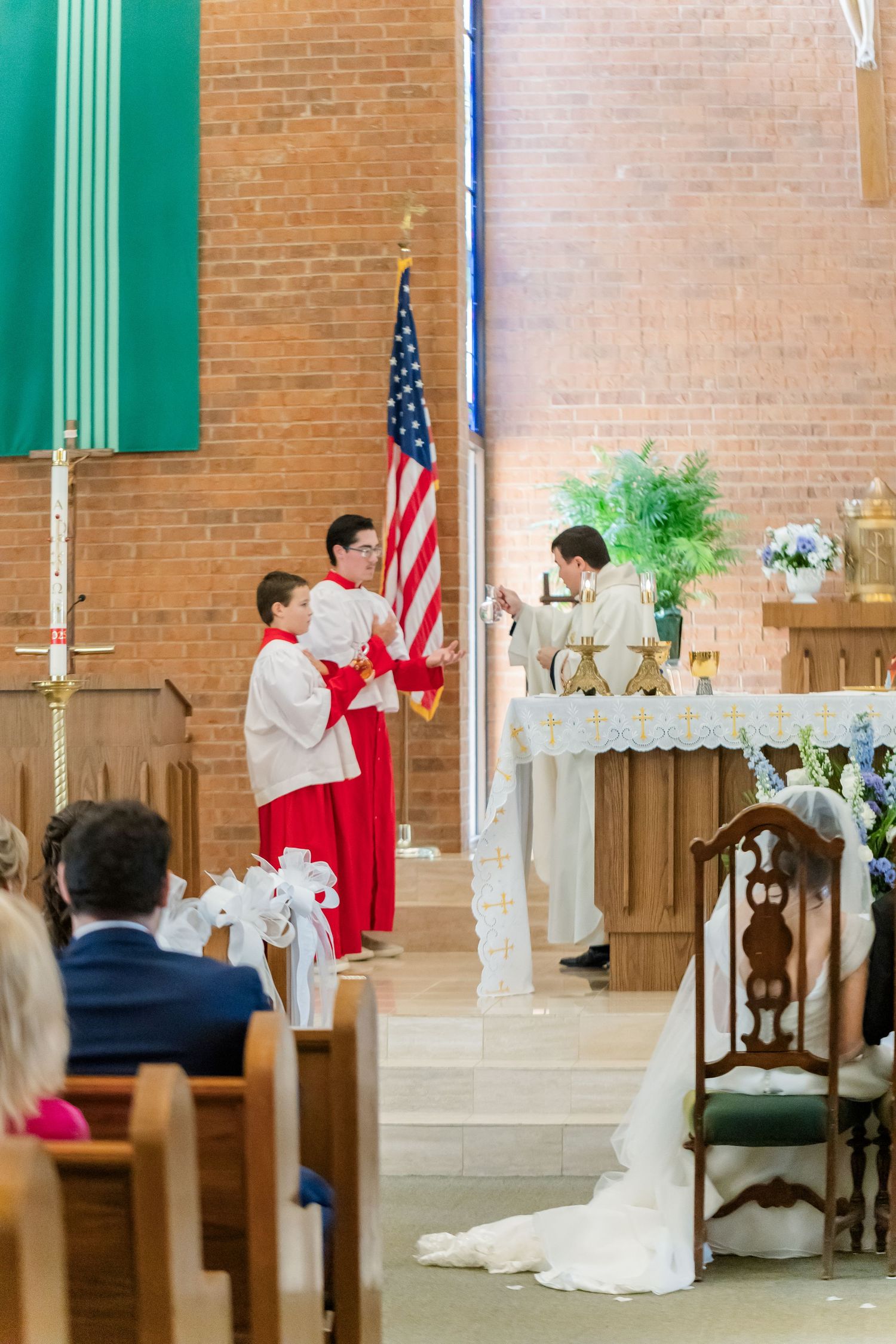 Catholic church wedding ceremony with white floral arrangements and American flag at brick altar.