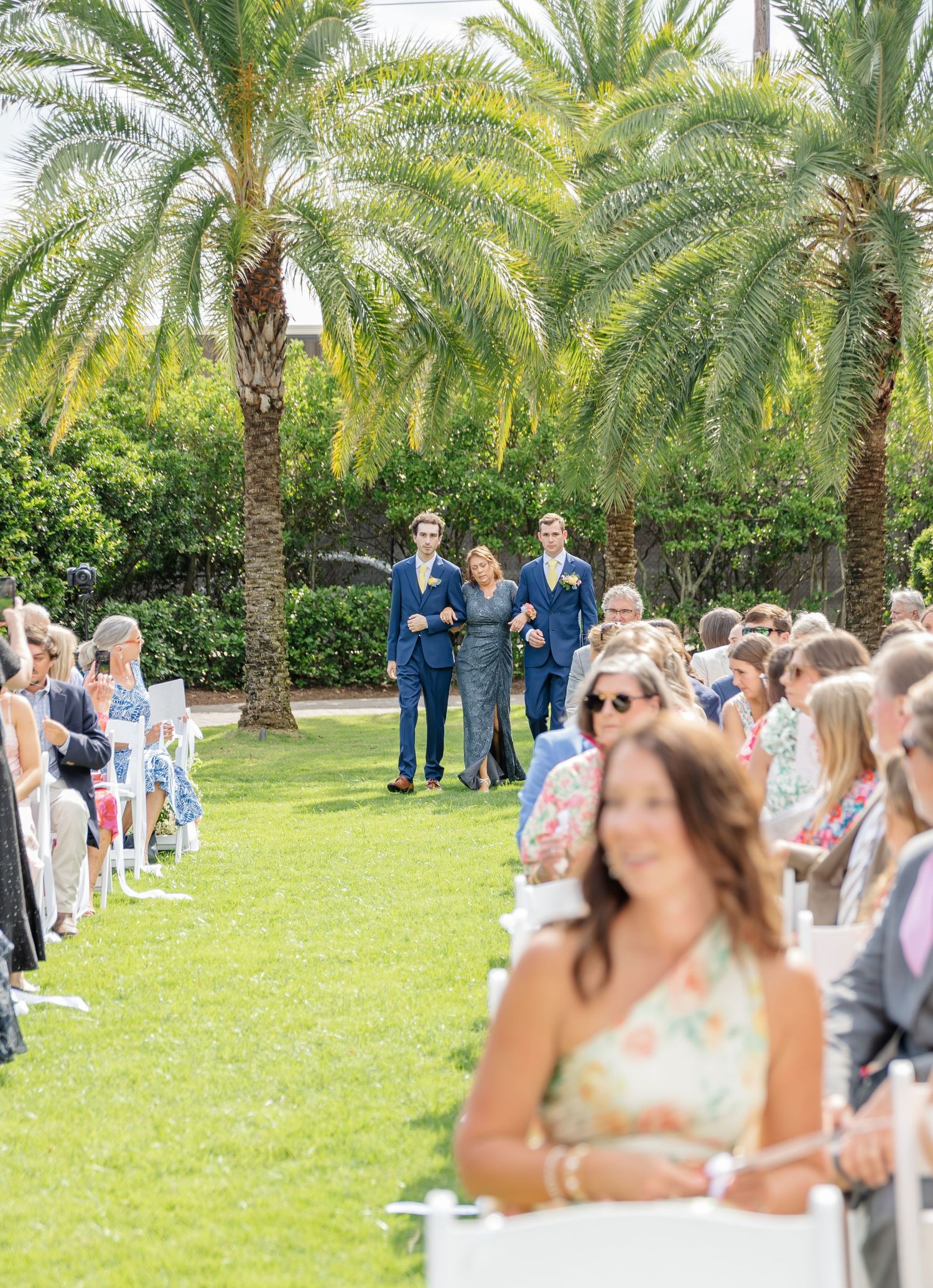 An outdoor wedding ceremony setup with palm trees and guests seated on white chairs on a manicured lawn.