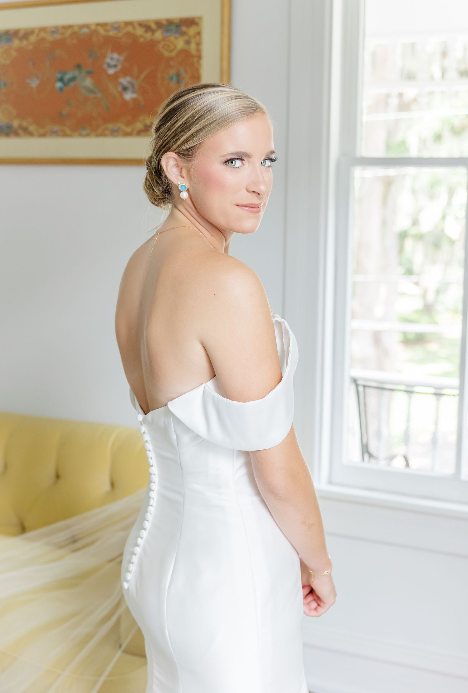 A bride in an elegant white off-shoulder wedding dress poses near a bright window in a classic interior setting.