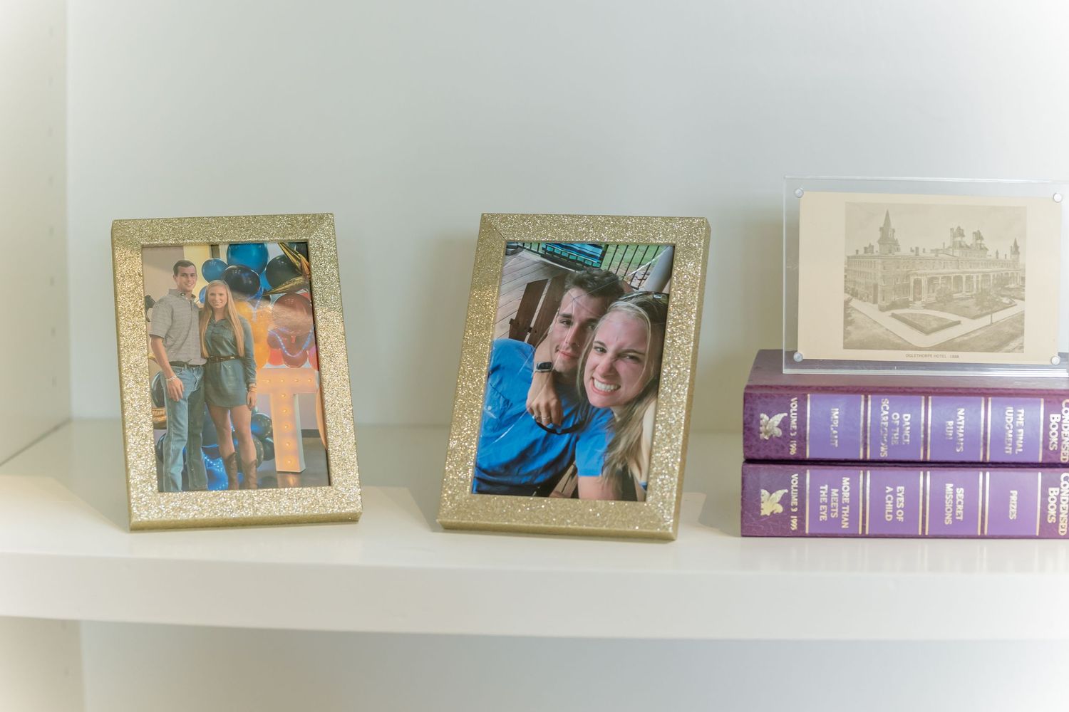 Framed photographs and purple books displayed on a white shelf against a light wall.