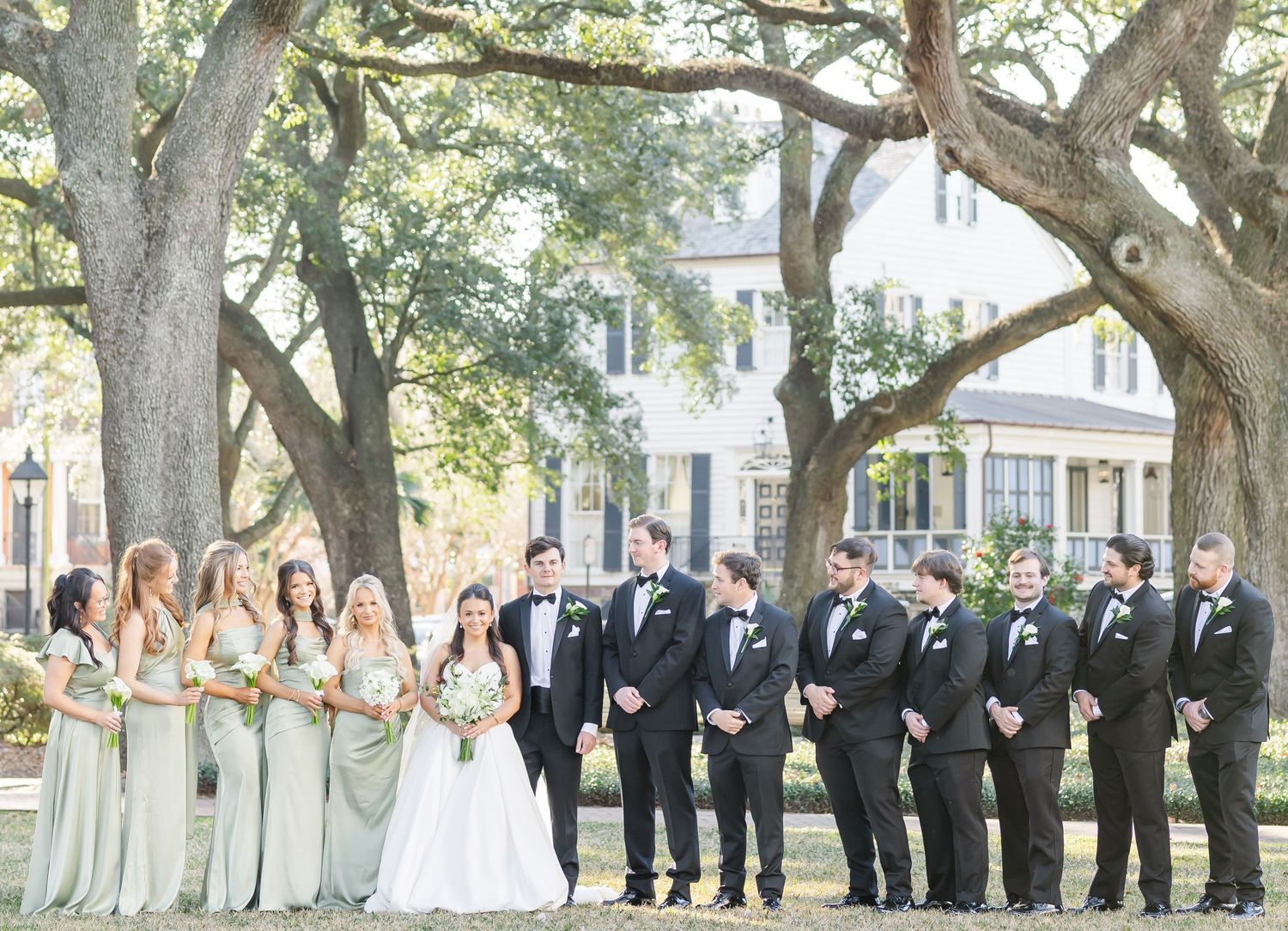 Wedding party lines up under oak trees wearing black tuxedos and mint green dresses.