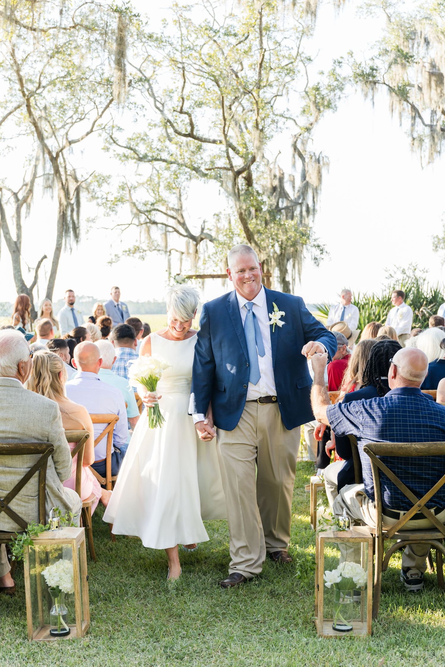 Wedding guests seated in white chairs under oak trees during an outdoor ceremony by the water.