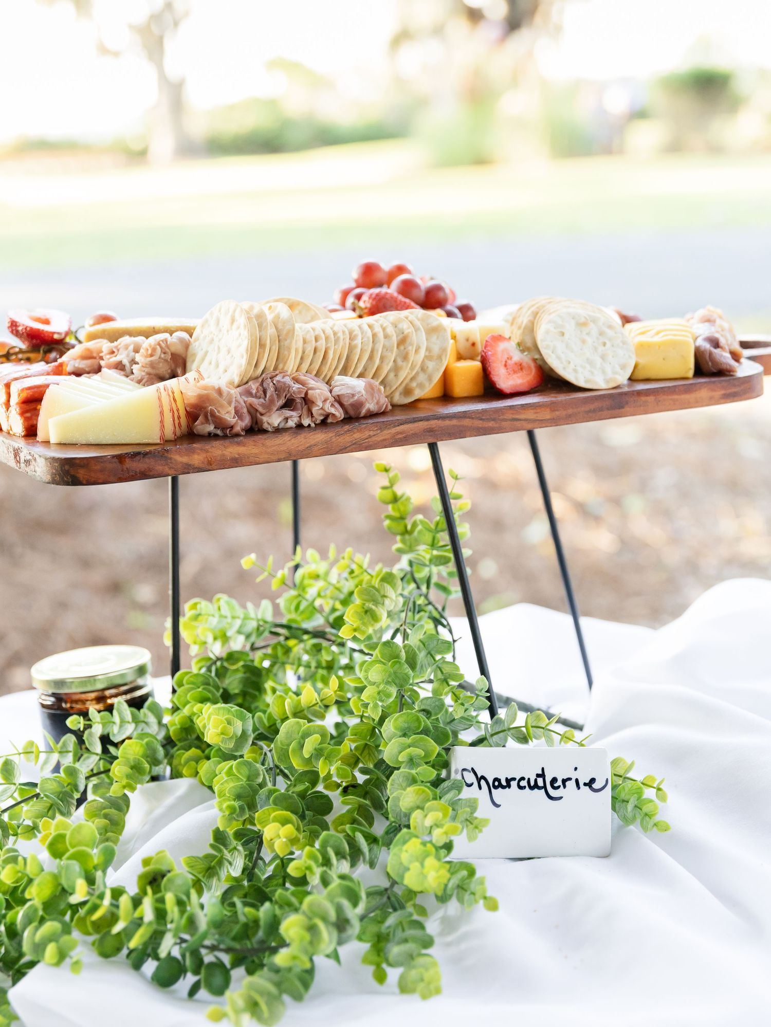 Long wooden charcuterie board with assorted meats, cheeses and garnishes displayed on white linens.