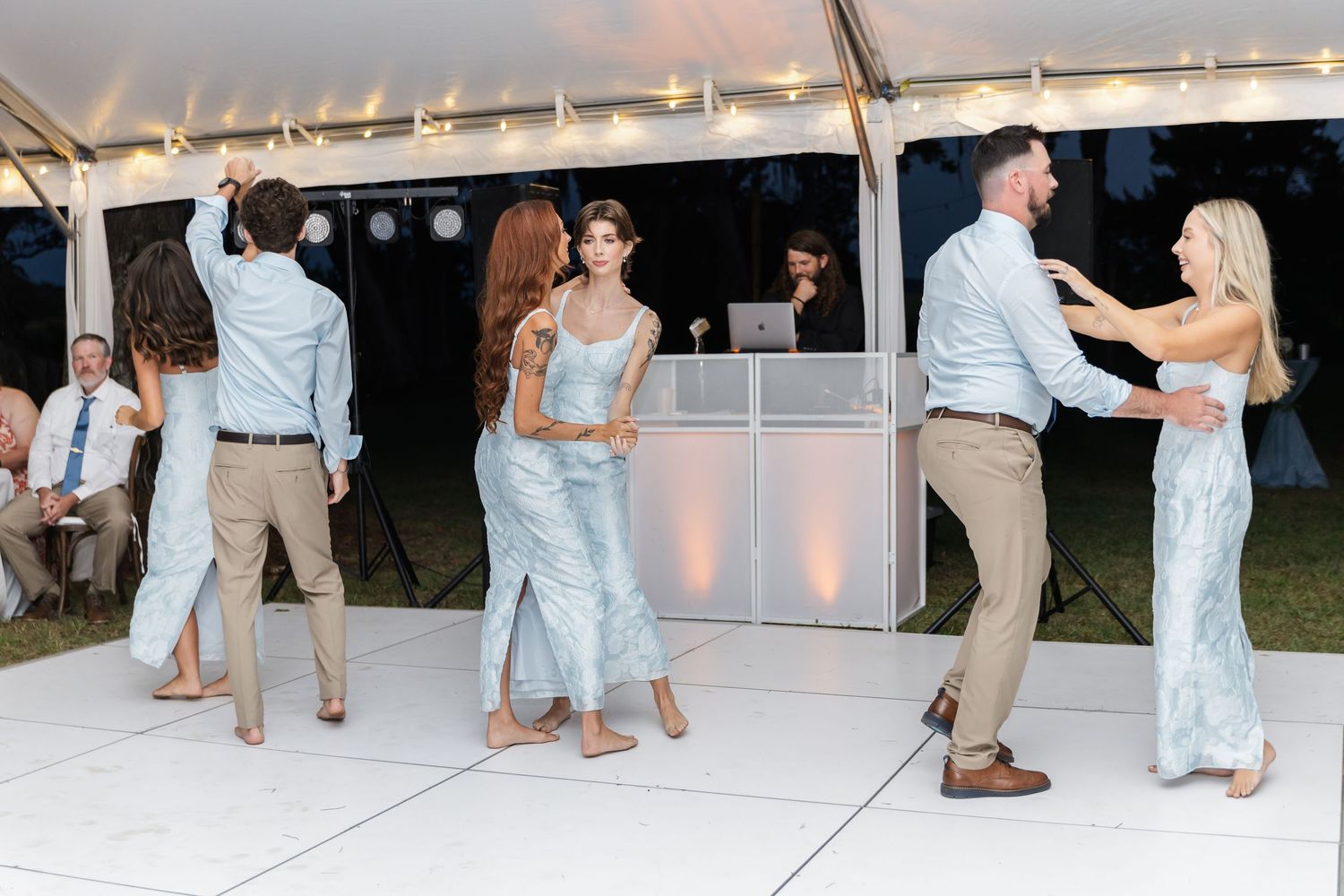 Wedding reception guests dancing on a white dance floor under a tent with string lights.
