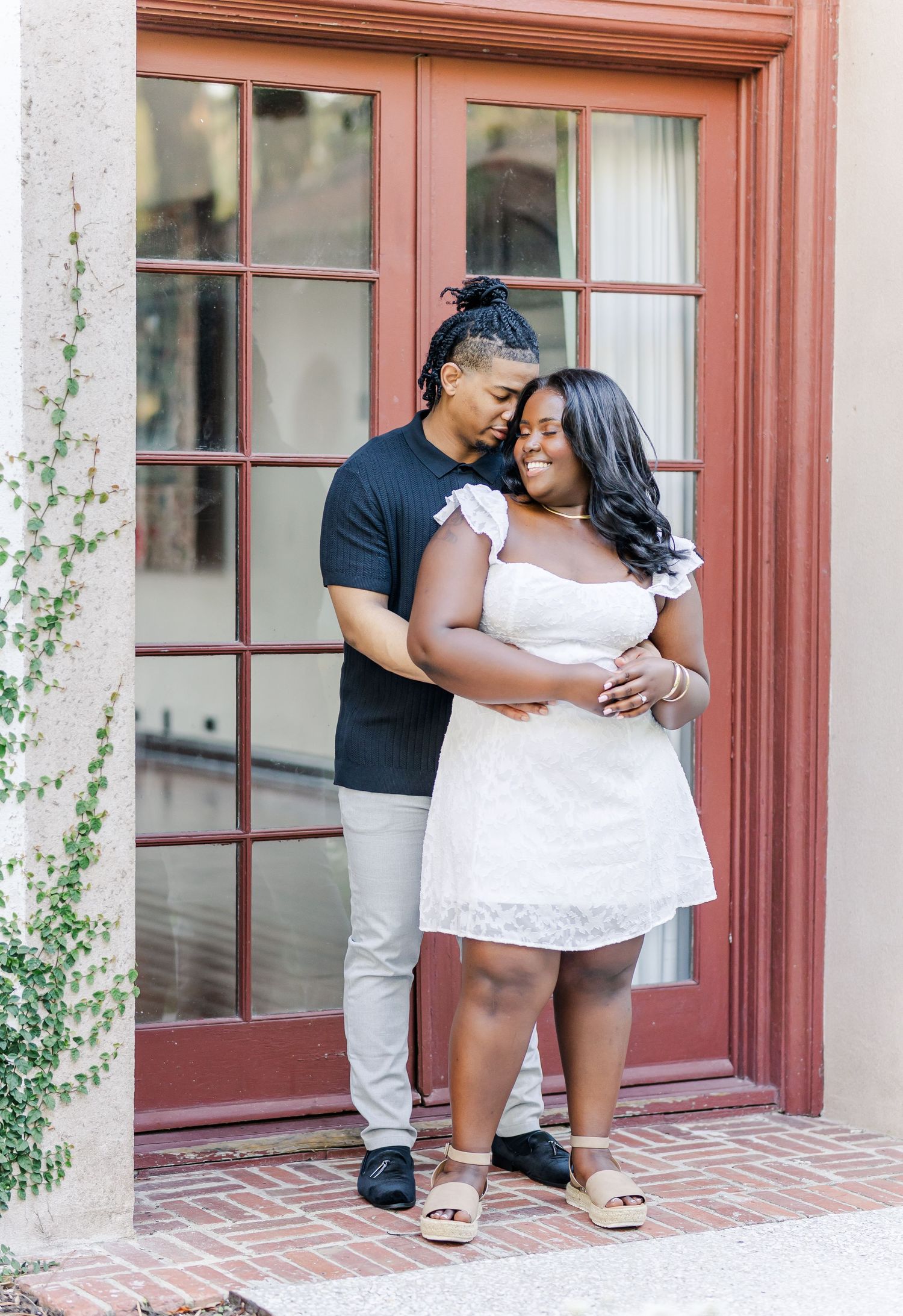 A couple embraces tenderly in front of a red French door, woman in white dress, man in black shirt.