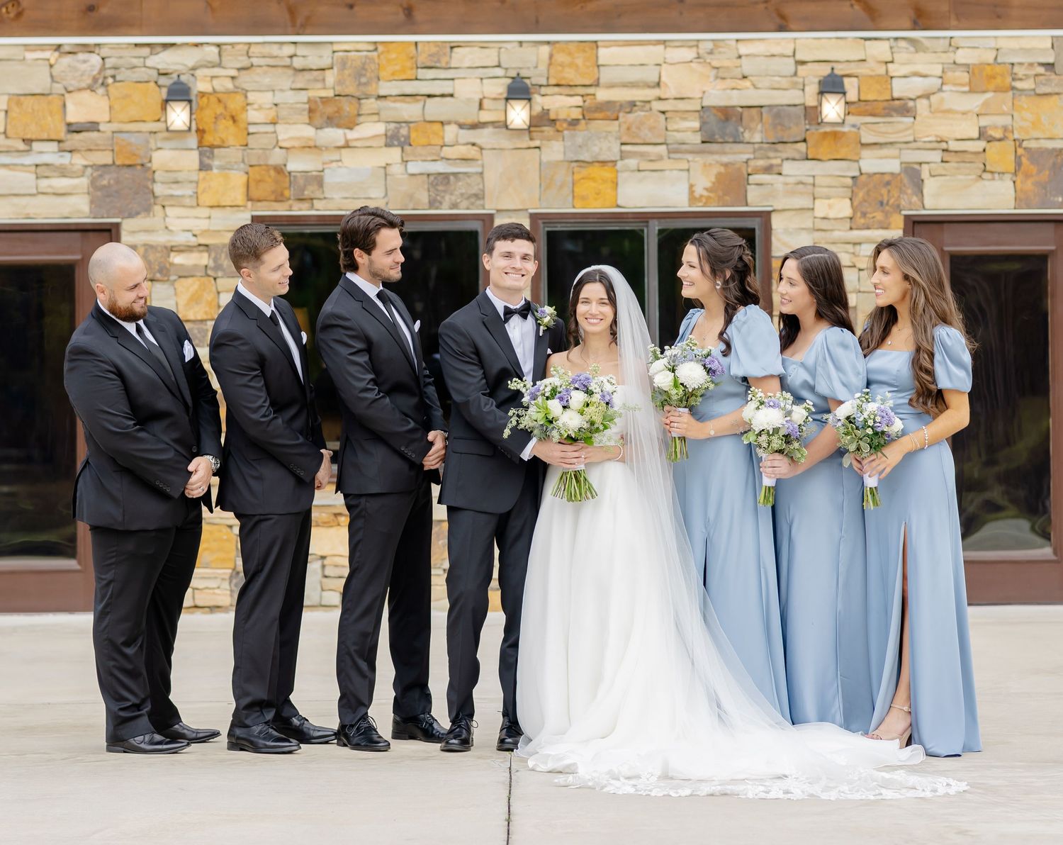 Wedding party lineup with bridesmaids in blue and groomsmen in black against stone backdrop.