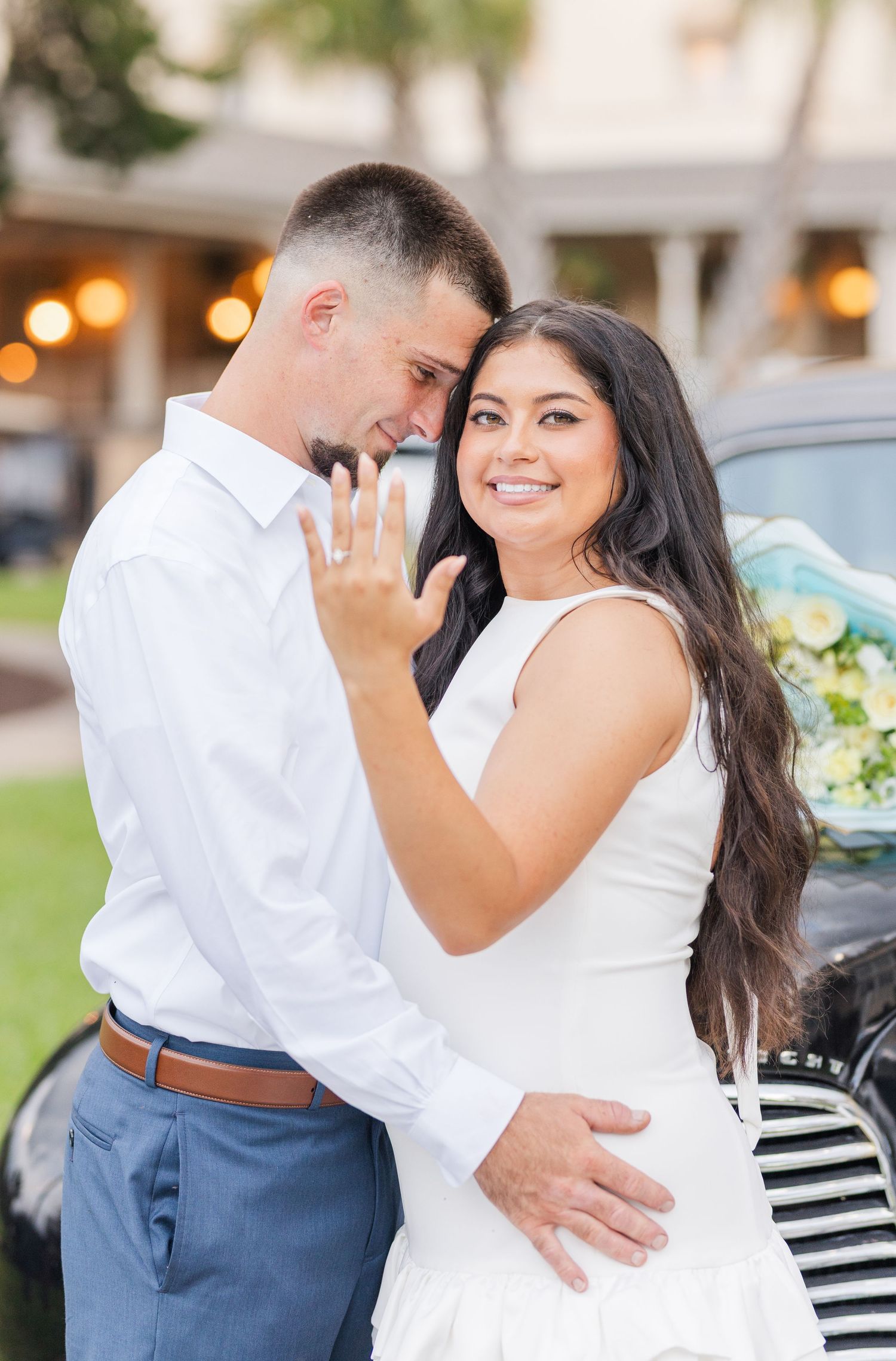 Smiling bride in white dress shows off engagement ring while embracing groom near a vintage black car.