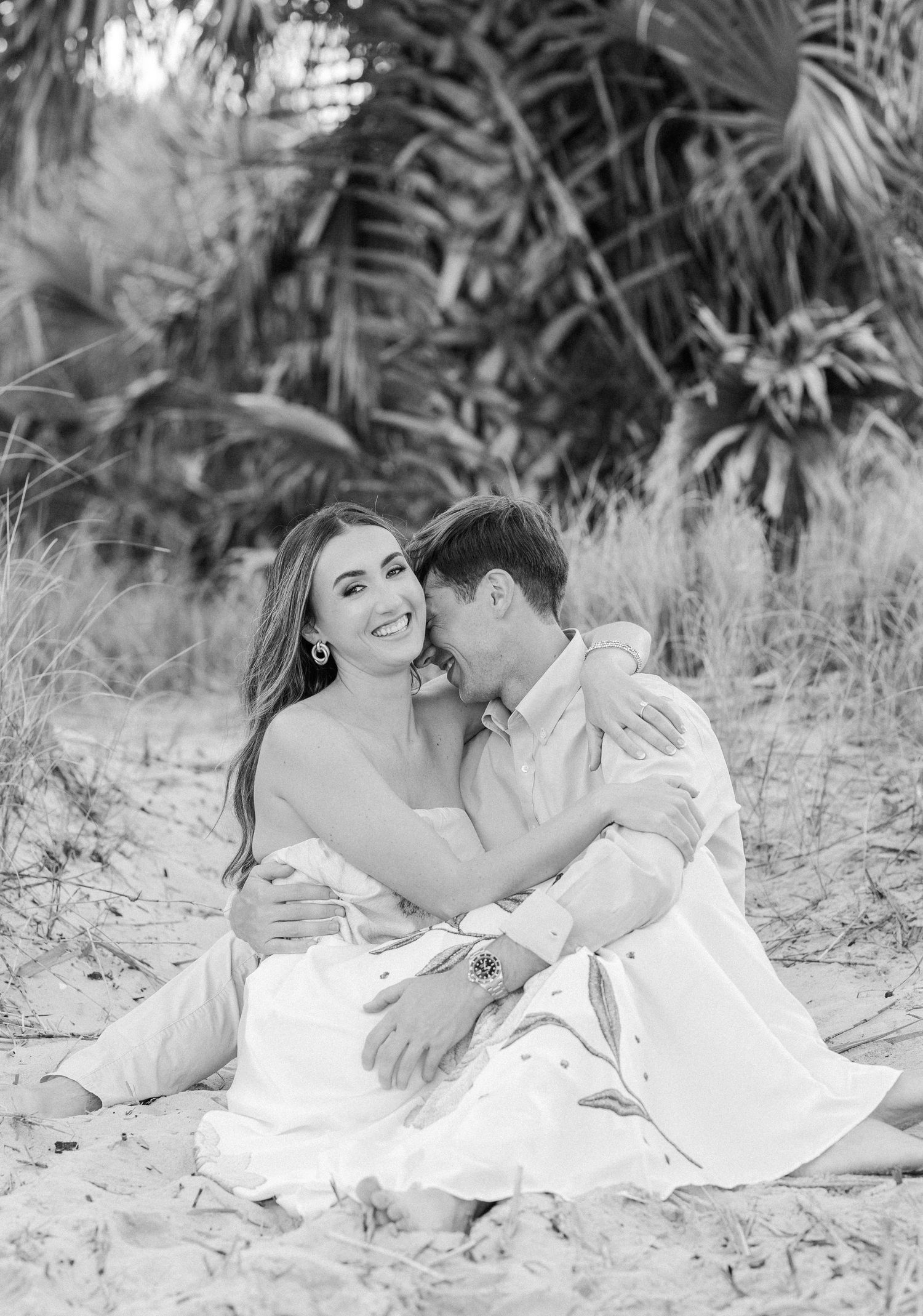 Black and white photo of couple sitting on beach sand embracing romantically near palm trees.