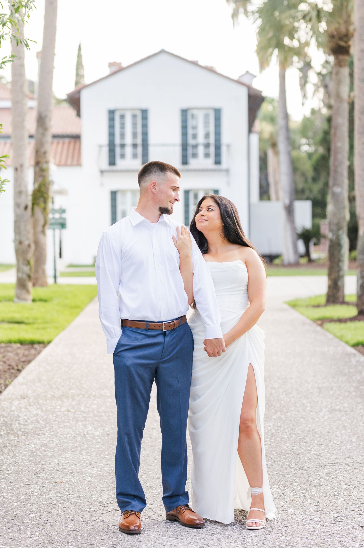 Couple in matching white outfits walking hand in hand on pathway in front of white building with palms.
