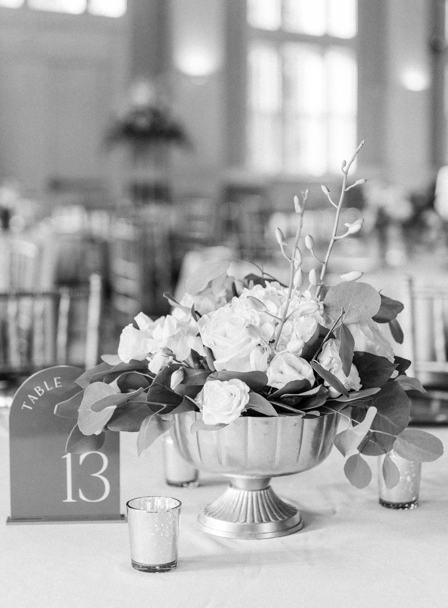 Black and white wedding reception centerpiece with white roses in silver compote vase beside table number 13 sign.