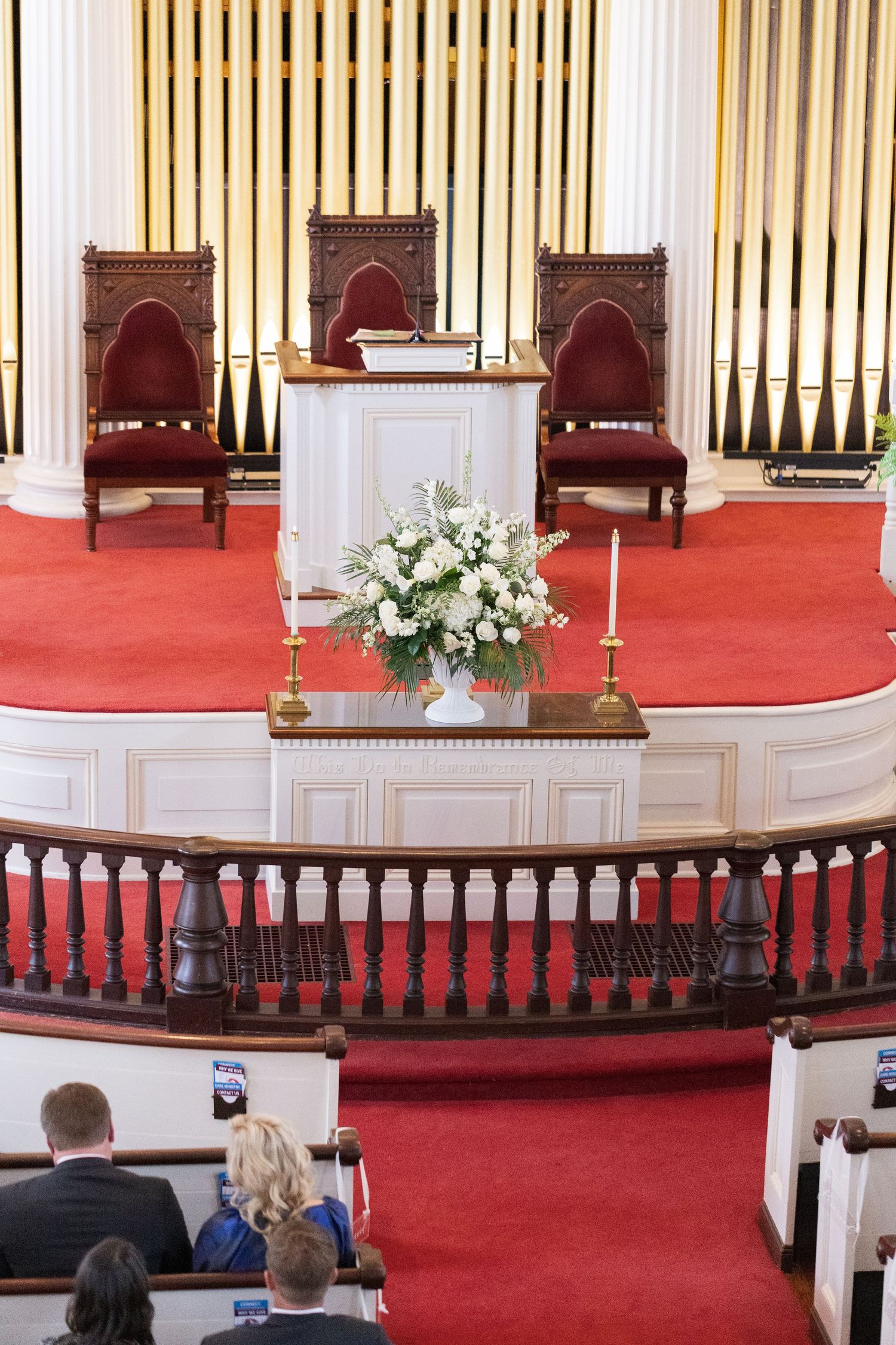 Elegant church altar with white pulpit, floral arrangement, candles, red carpet, and ornate wooden chairs on a raised platform.