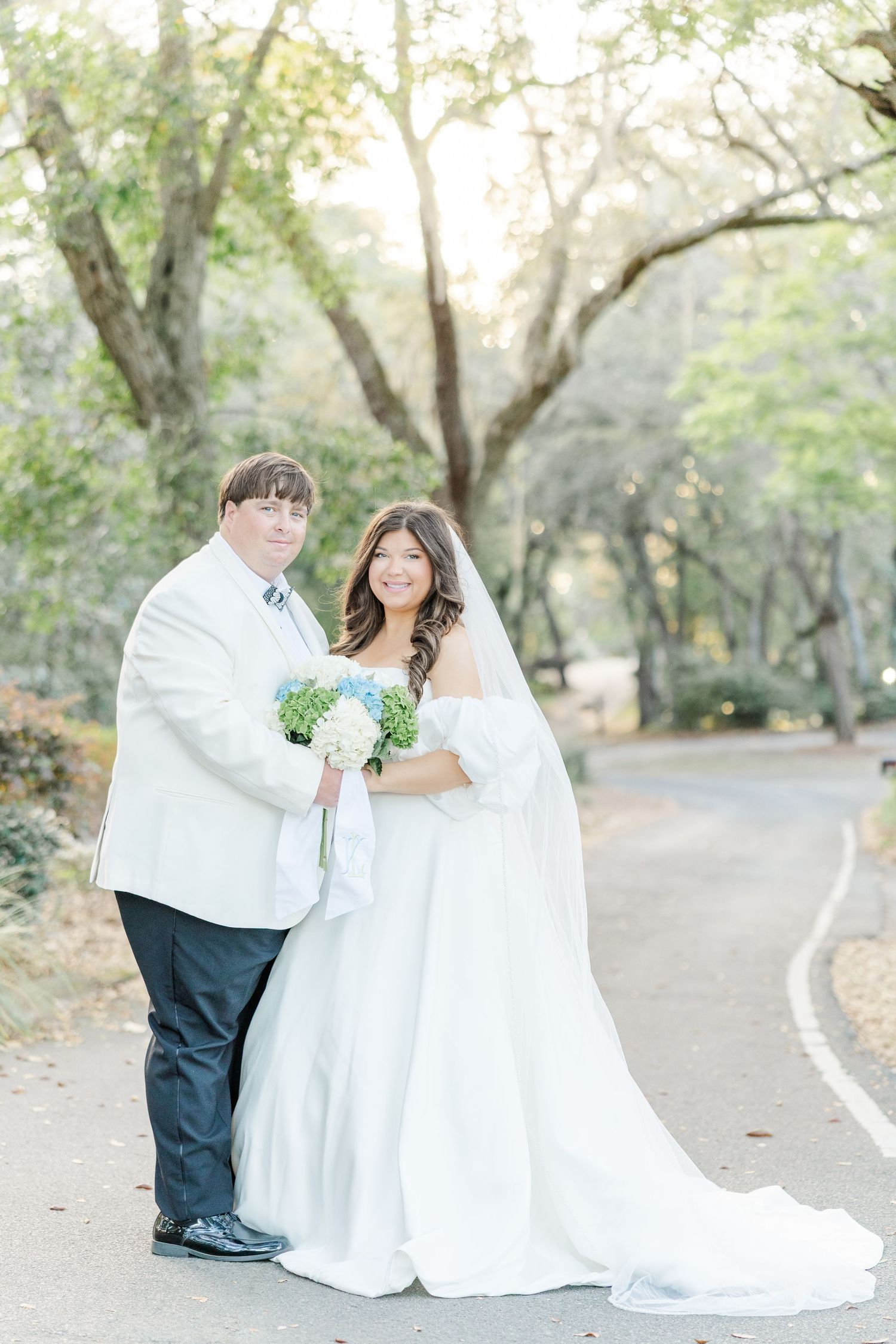 Bride and groom posing on a tree-lined path, bride in white gown with veil holding blue hydrangea bouquet.