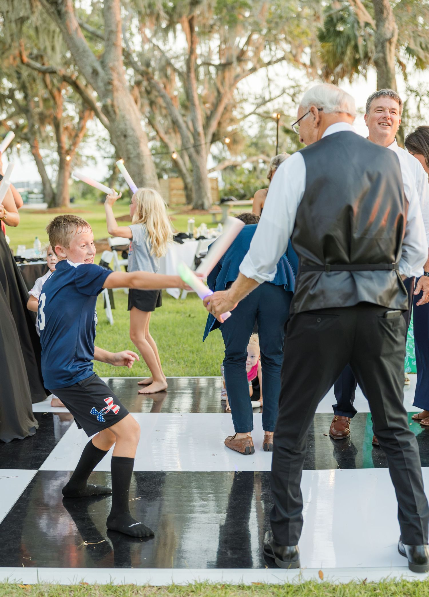 A young boy energetically dances with an elderly man in a vest at an outdoor wedding reception on a black and white dance floor.