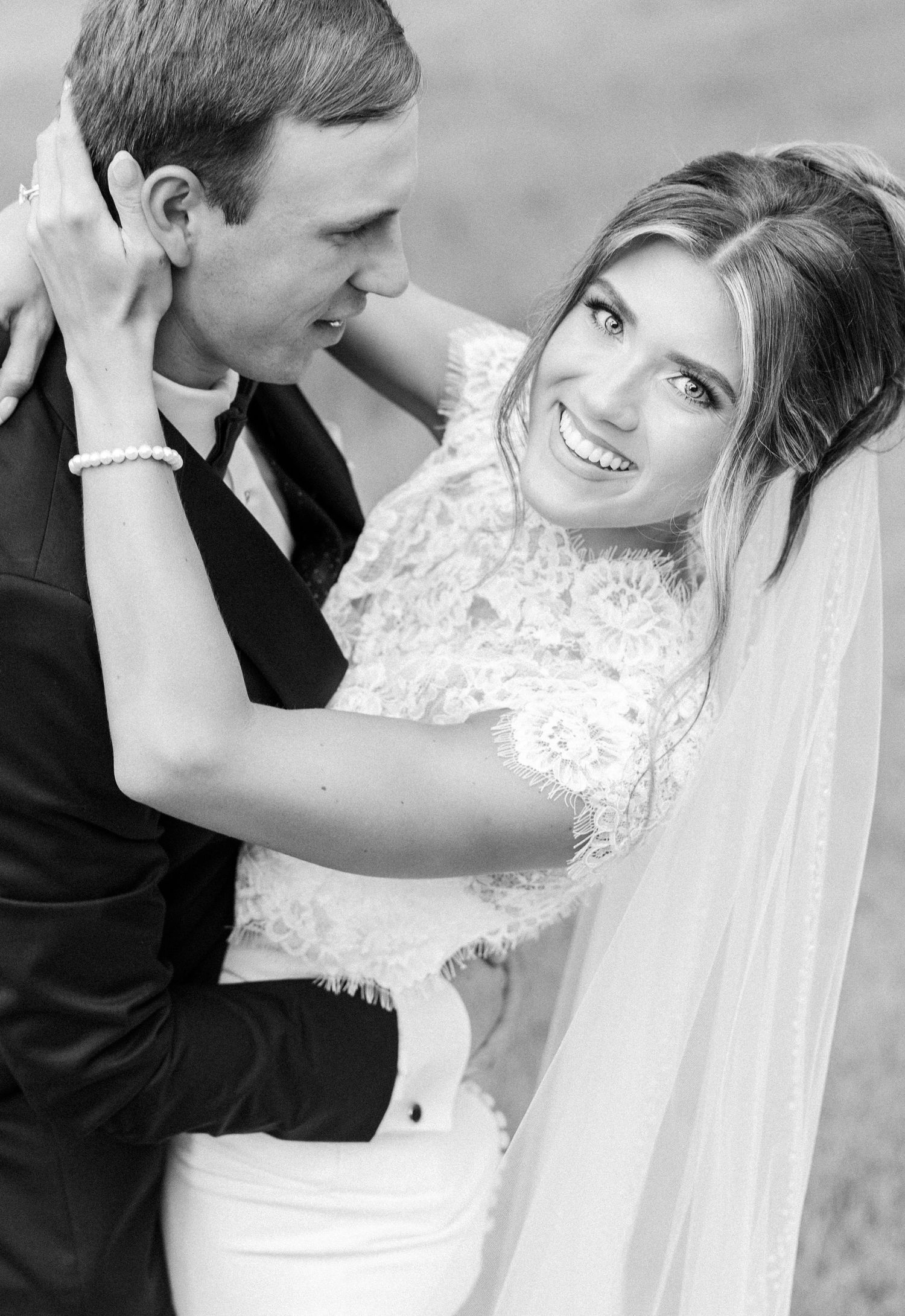 Black and white portrait of smiling bride in lace dress embracing groom at outdoor wedding.