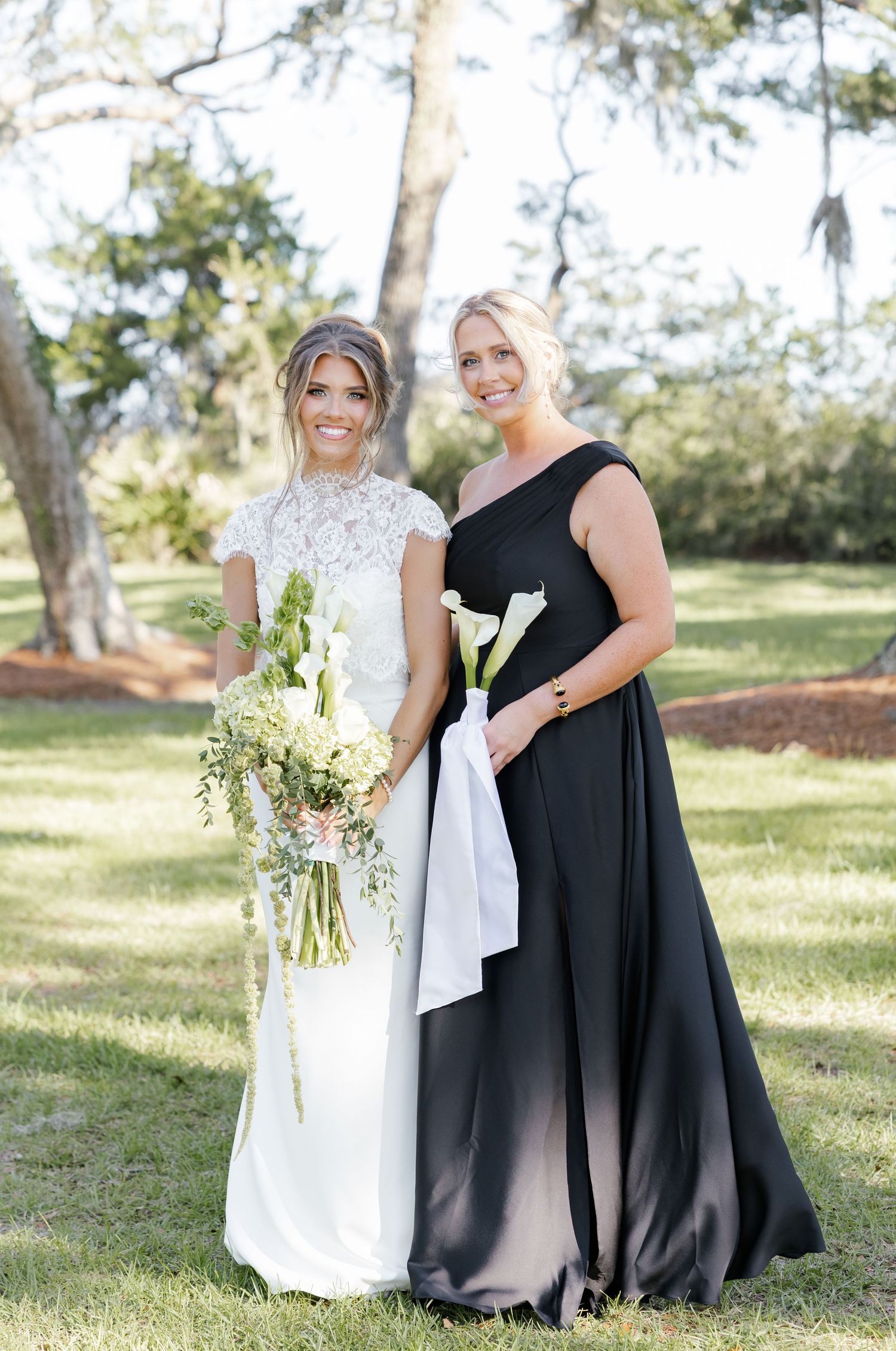 Bride in white lace gown holds bouquet beside bridesmaid in black ombre one-shoulder dress outdoors.