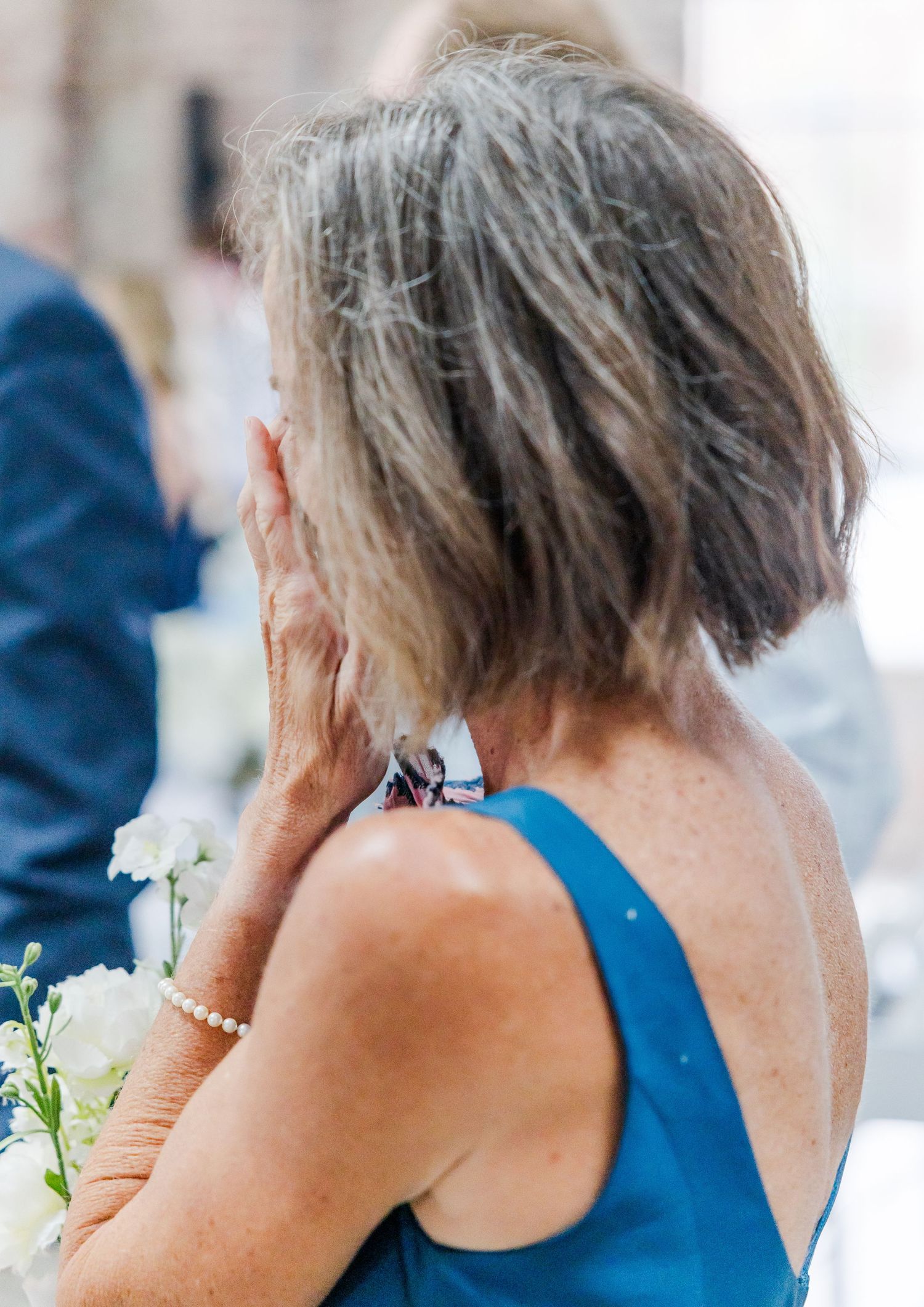 Emotional wedding guest in teal dress wipes tears, holding white flowers during ceremony.