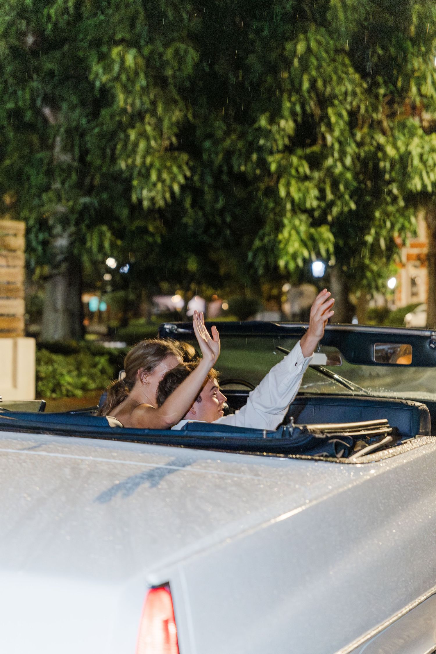 Bride waving goodbye from classic convertible car surrounded by lush green trees after wedding reception.