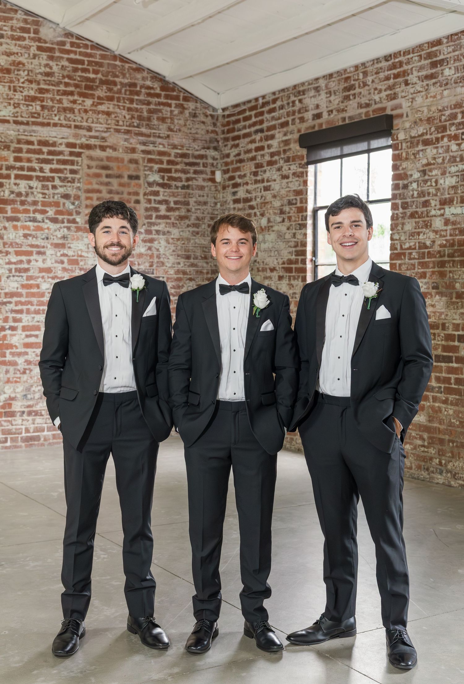 Three groomsmen in black tuxedos with bow ties and white boutonnieres pose in a brick venue.