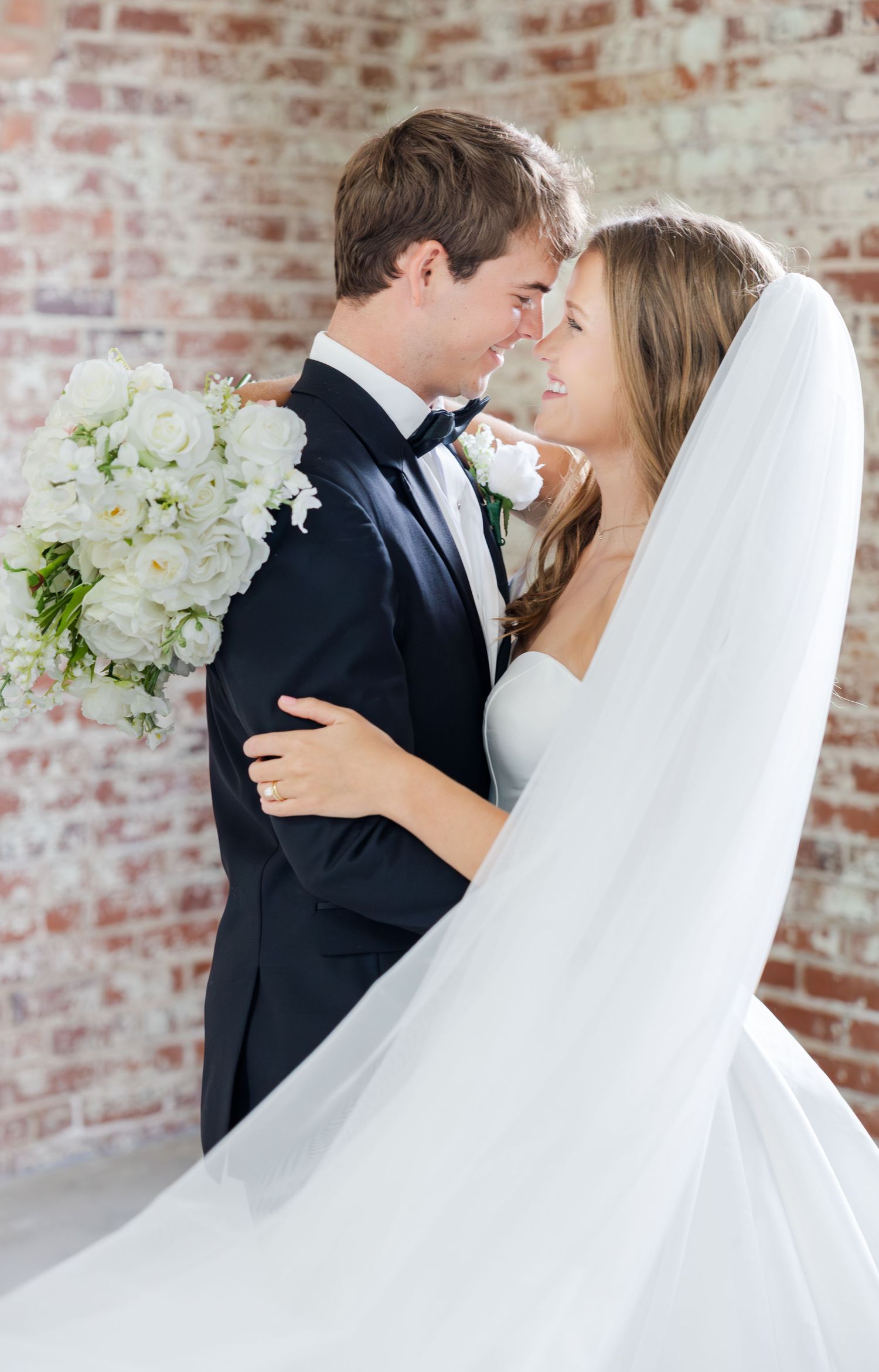 Bride and groom share an intimate forehead-to-forehead moment, bride holding white floral bouquet against brick wall.