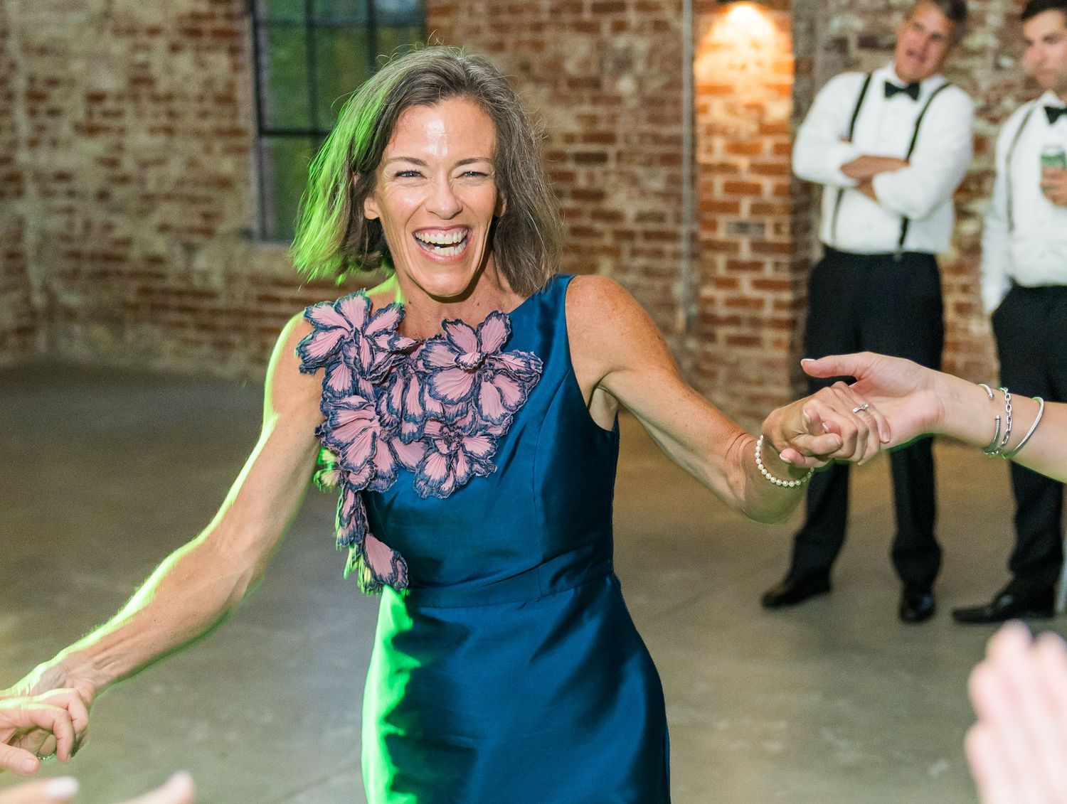 Woman in blue floral dress laughs joyfully while dancing at wedding reception in brick-walled venue.