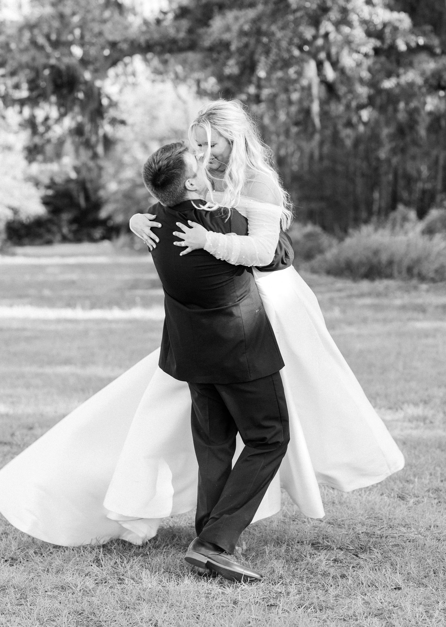 Black and white photo of groom lifting bride in wedding dress outdoors under tree canopy.