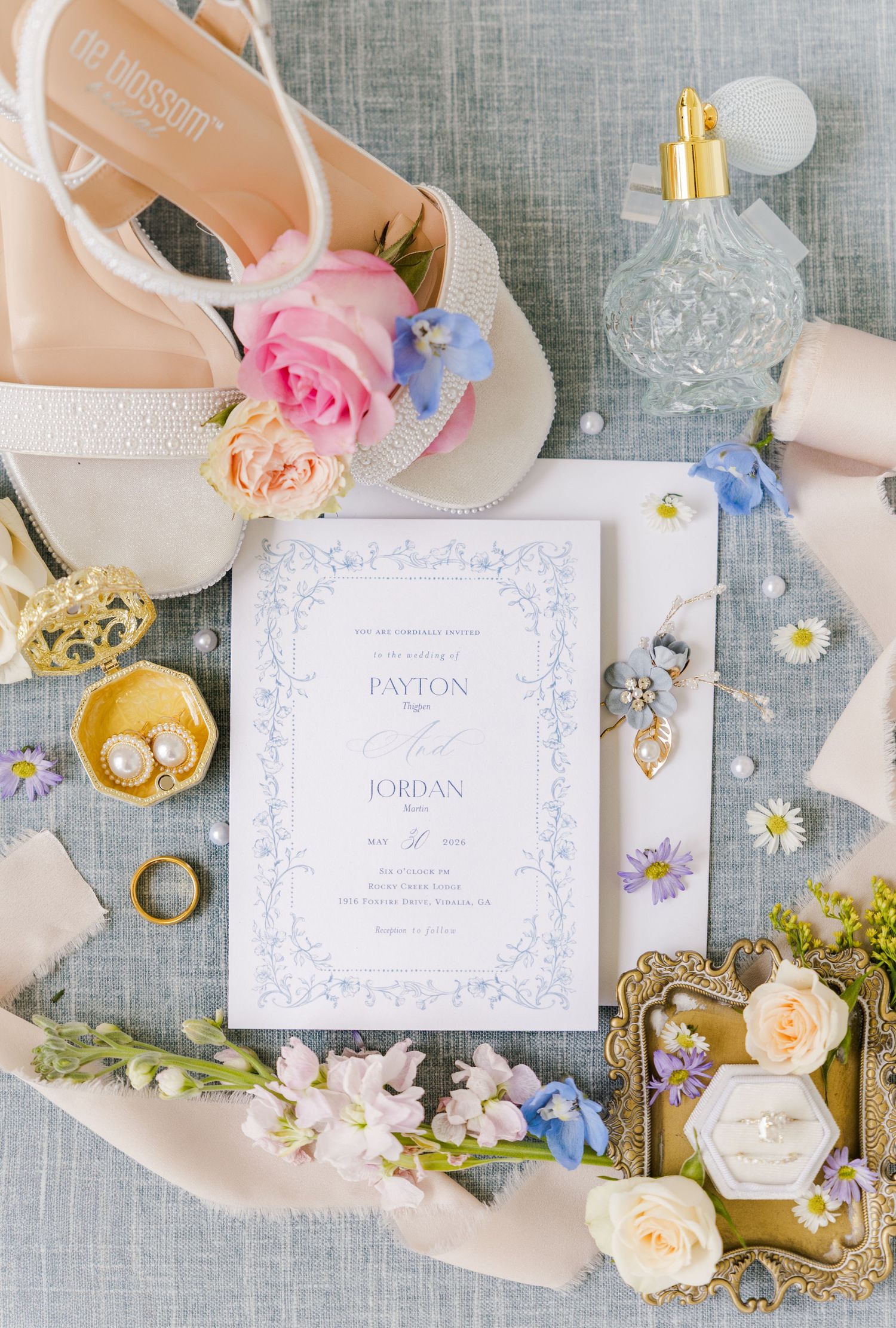 Elegant wedding flat lay with blue floral invitation, bridal shoes, rings, and flowers on a gray linen background.