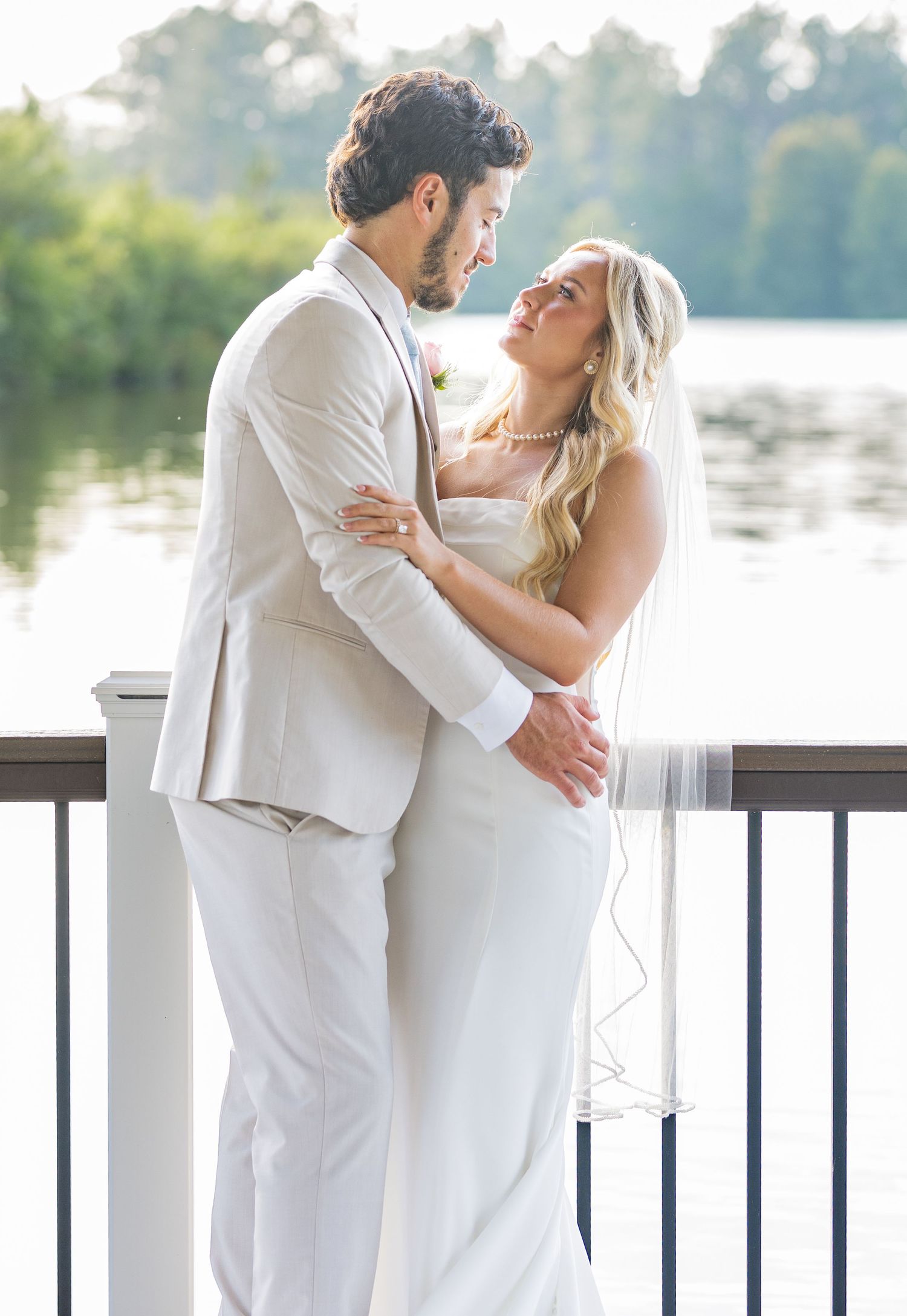 Bride and groom share a romantic embrace on a waterfront deck, with the bride in a white gown and veil.