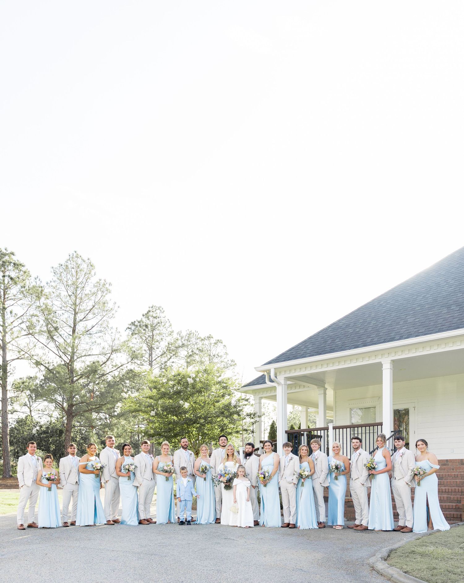 Large wedding party in light blue and beige attire posing outside white venue building.
