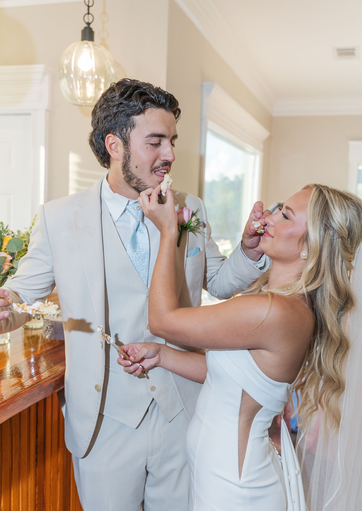 Bride and groom in light gray suit playfully feed each other wedding cake indoors at reception.
