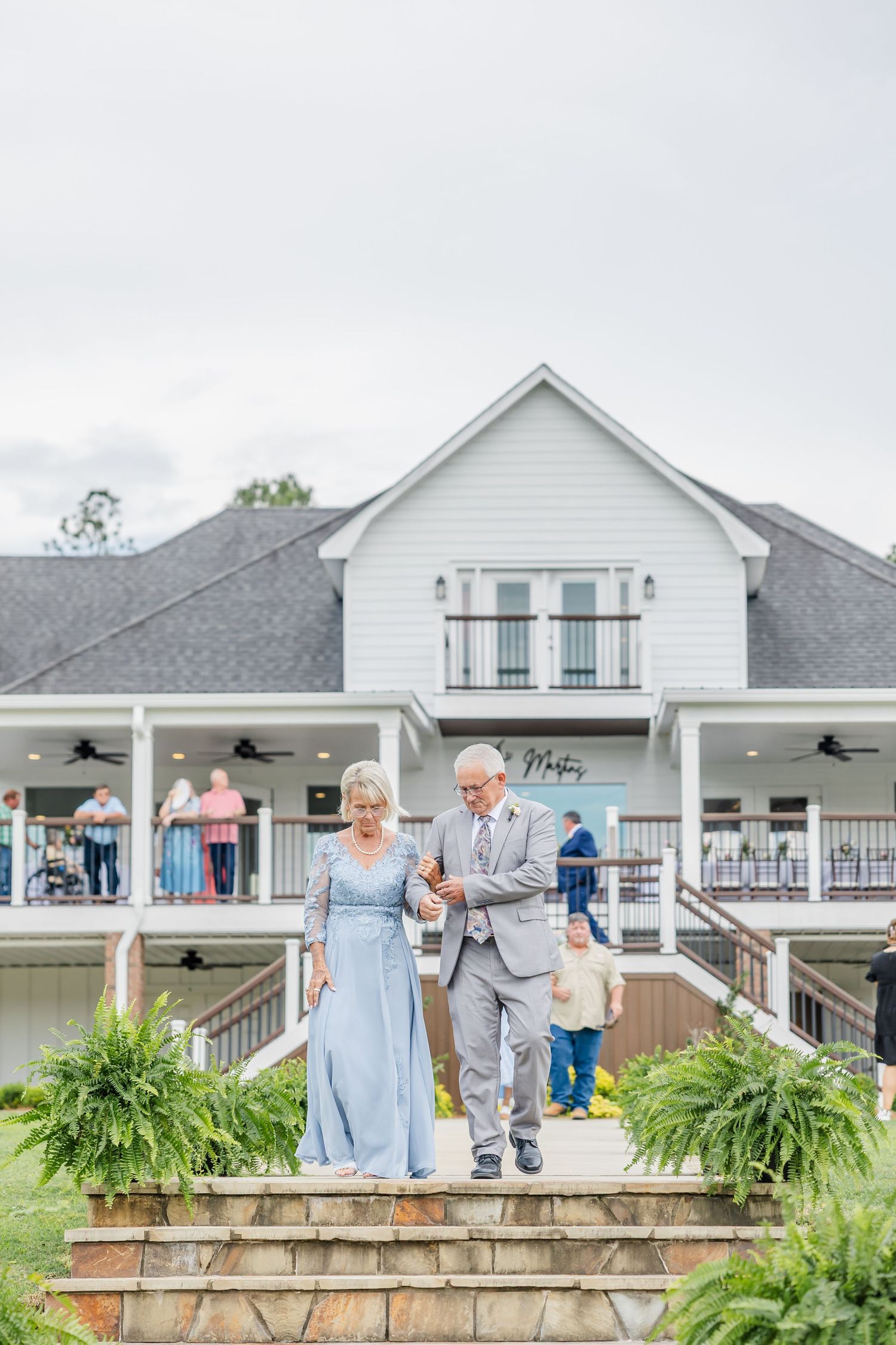 Elegant couple descends stone steps at white farmhouse wedding venue with guests watching from balcony.