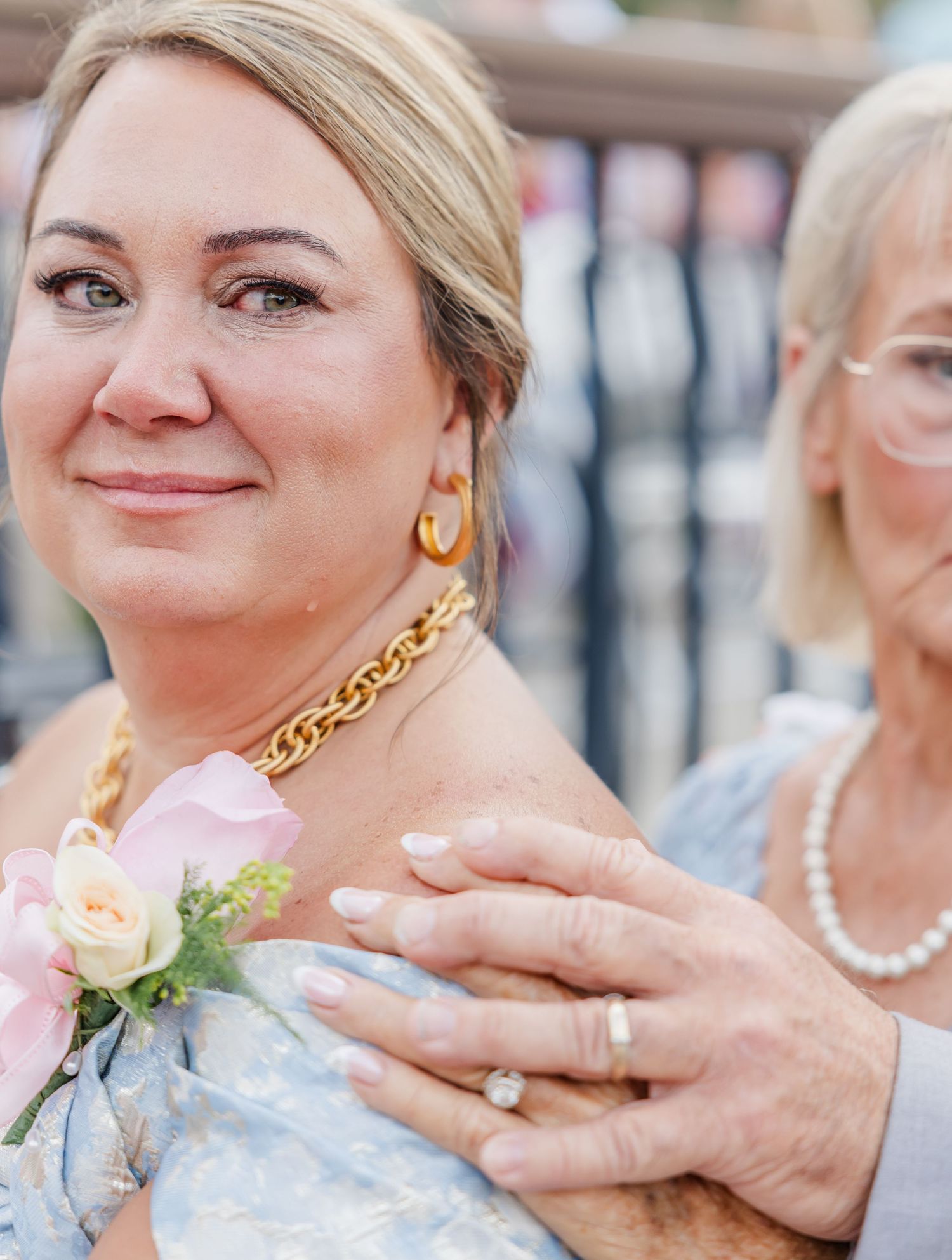 Close-up of a woman in light blue dress with gold jewelry and a pink rose boutonniere at a wedding.