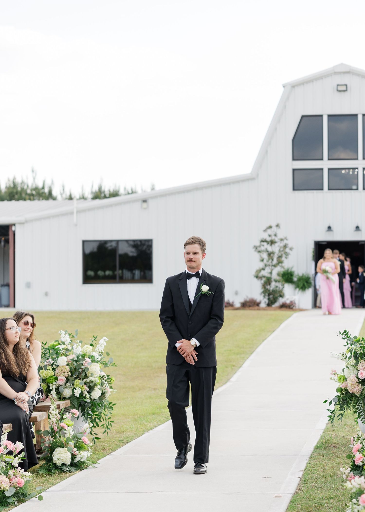 Groom in black tuxedo walks down outdoor wedding aisle flanked by floral arrangements and seated guests.