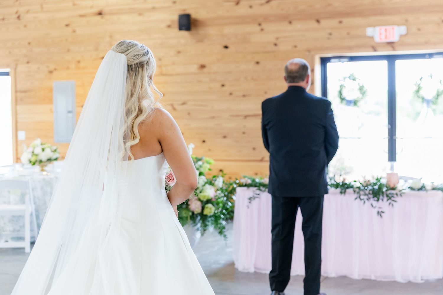 Bride in white gown and veil stands with father for first look moment inside rustic wedding venue.
