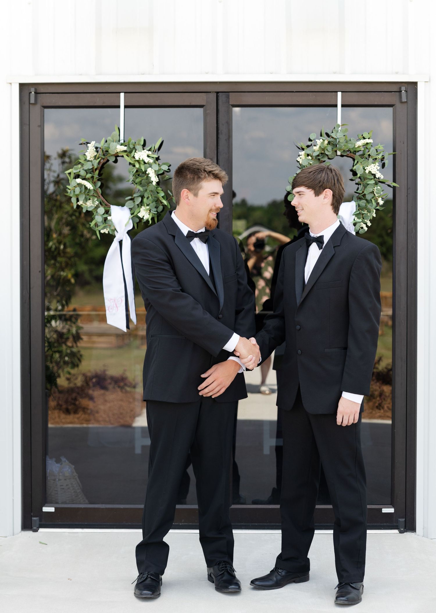 Two groomsmen in black tuxedos shaking hands before a white barn wedding venue decorated with floral wreaths.