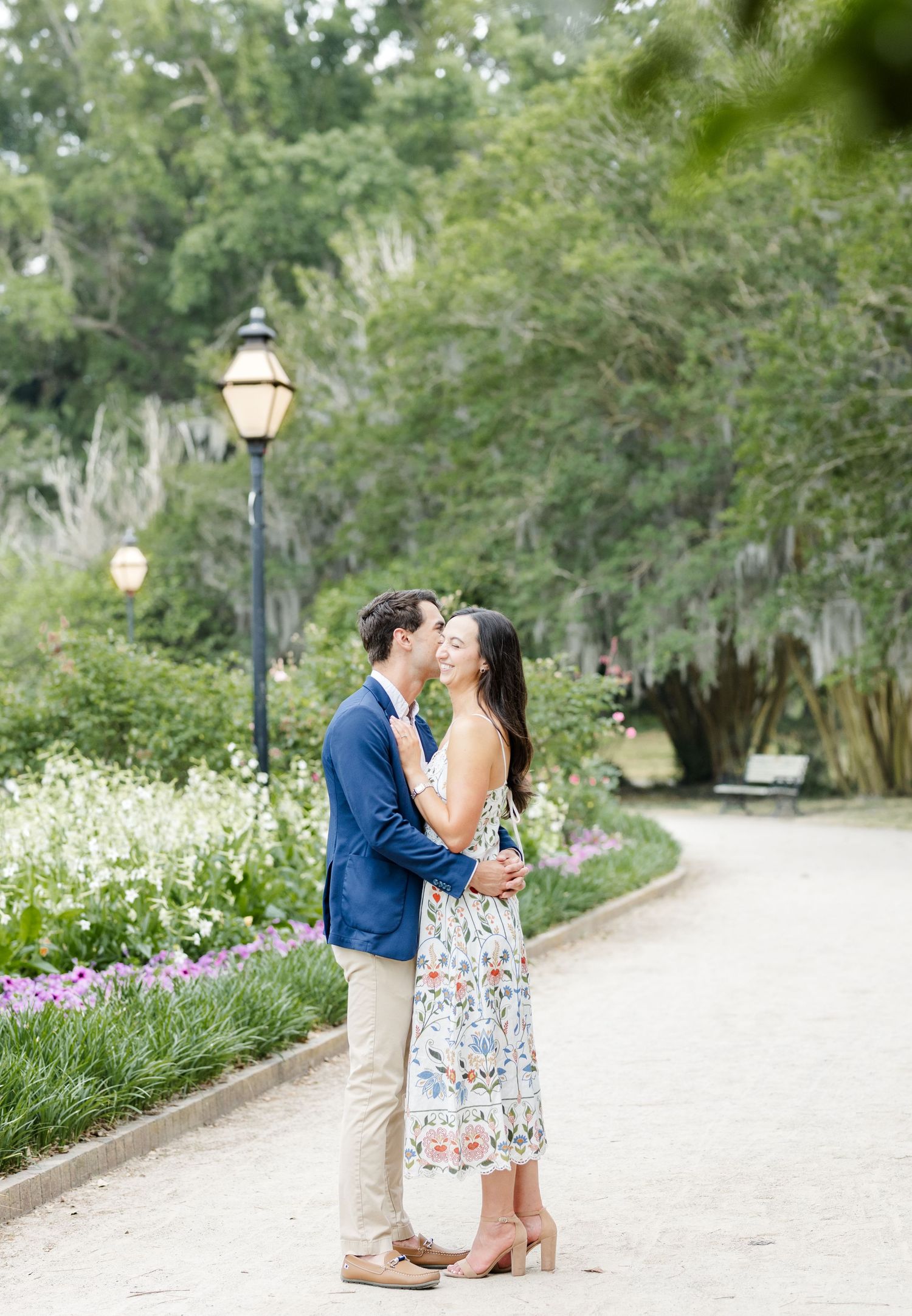 Couple sharing a romantic kiss in a lush garden pathway with blooming flowers and vintage lamp posts.