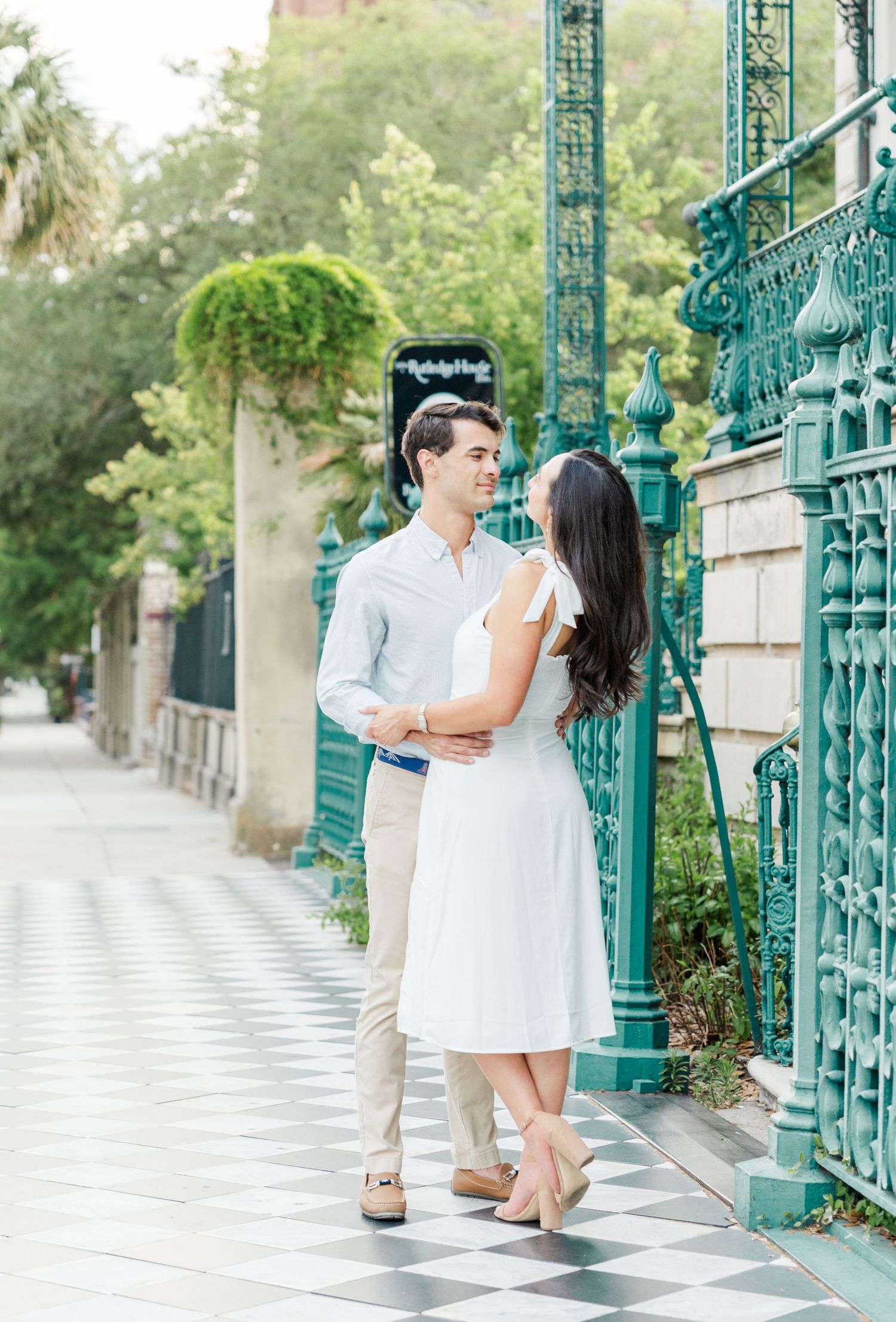 Couple in white outfits smiling at each other by ornate teal iron gates on a charming cobblestone sidewalk.