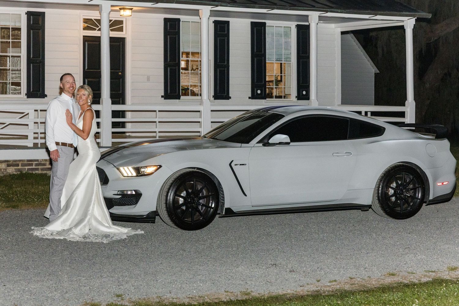 Bride and groom posing beside a white Ford Mustang Shelby GT350 in front of a historic white building.