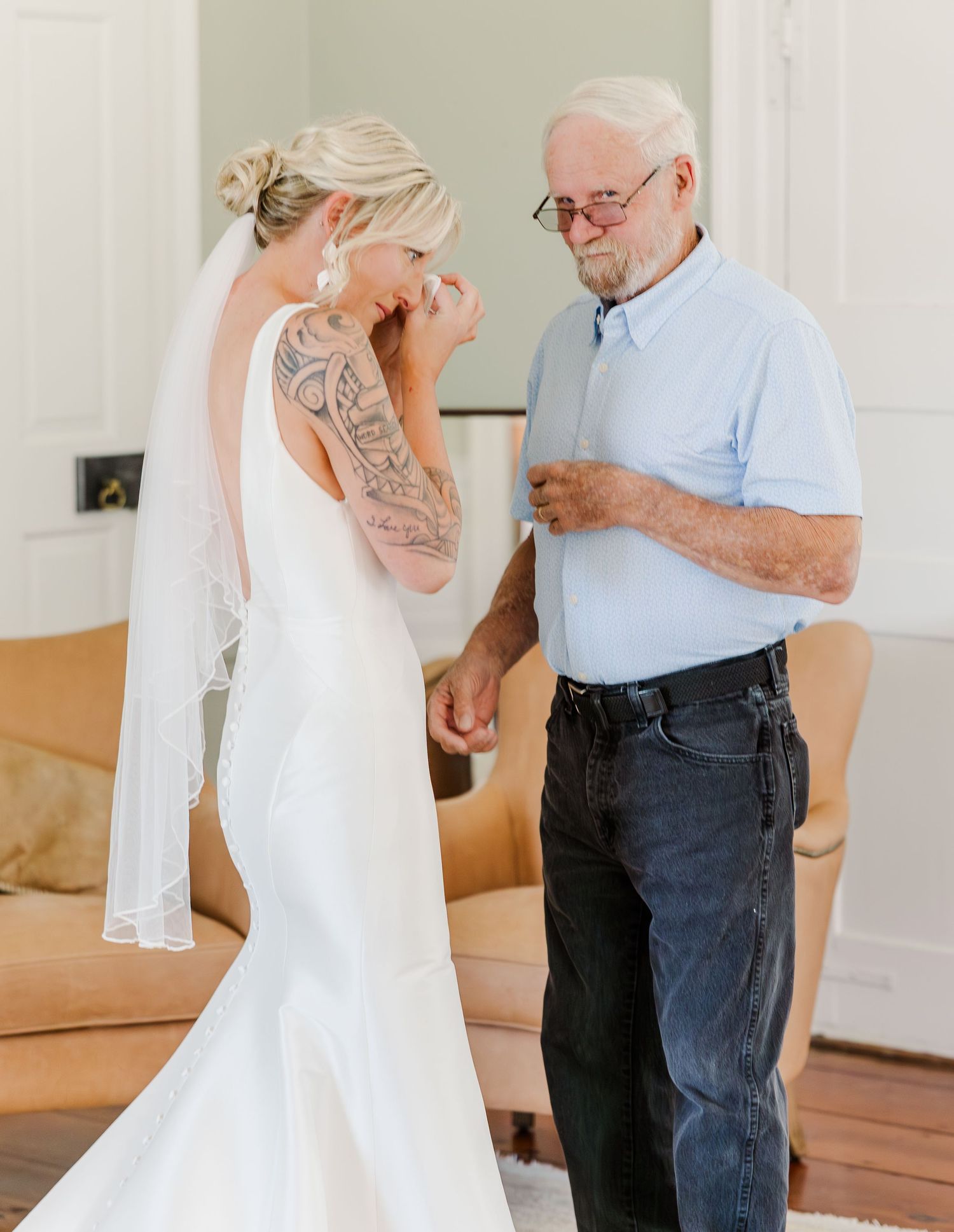 Bride in white mermaid gown with sleeve tattoo shares emotional moment with elderly man in blue shirt.