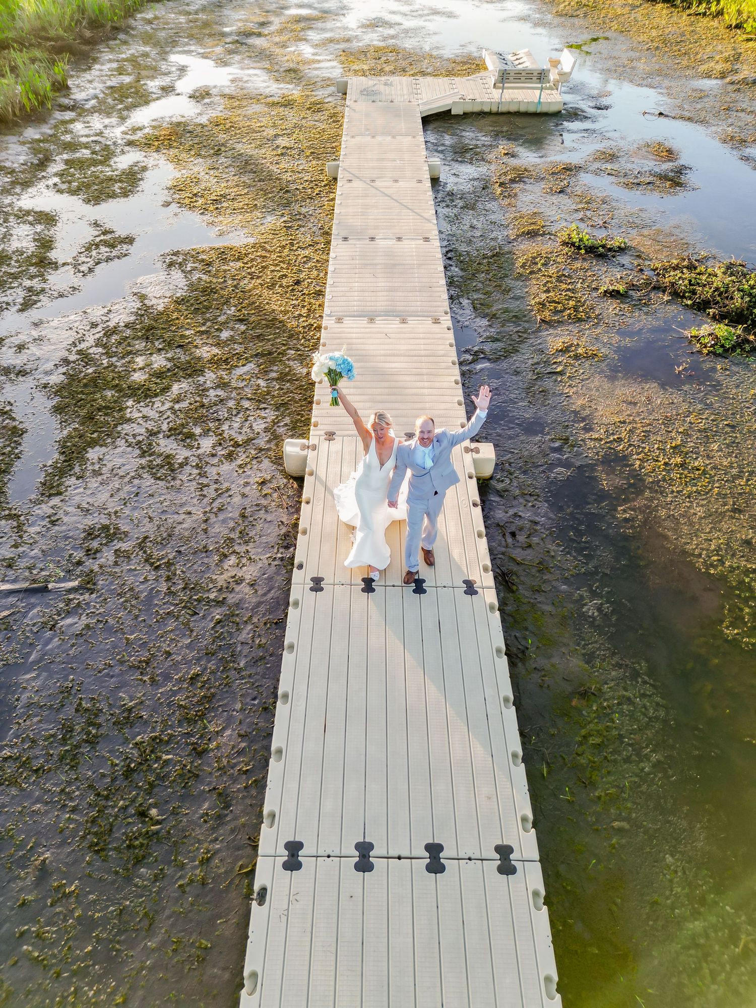 Aerial drone view of joyful bride and groom celebrating on wooden boardwalk dock surrounded by marsh wetlands.