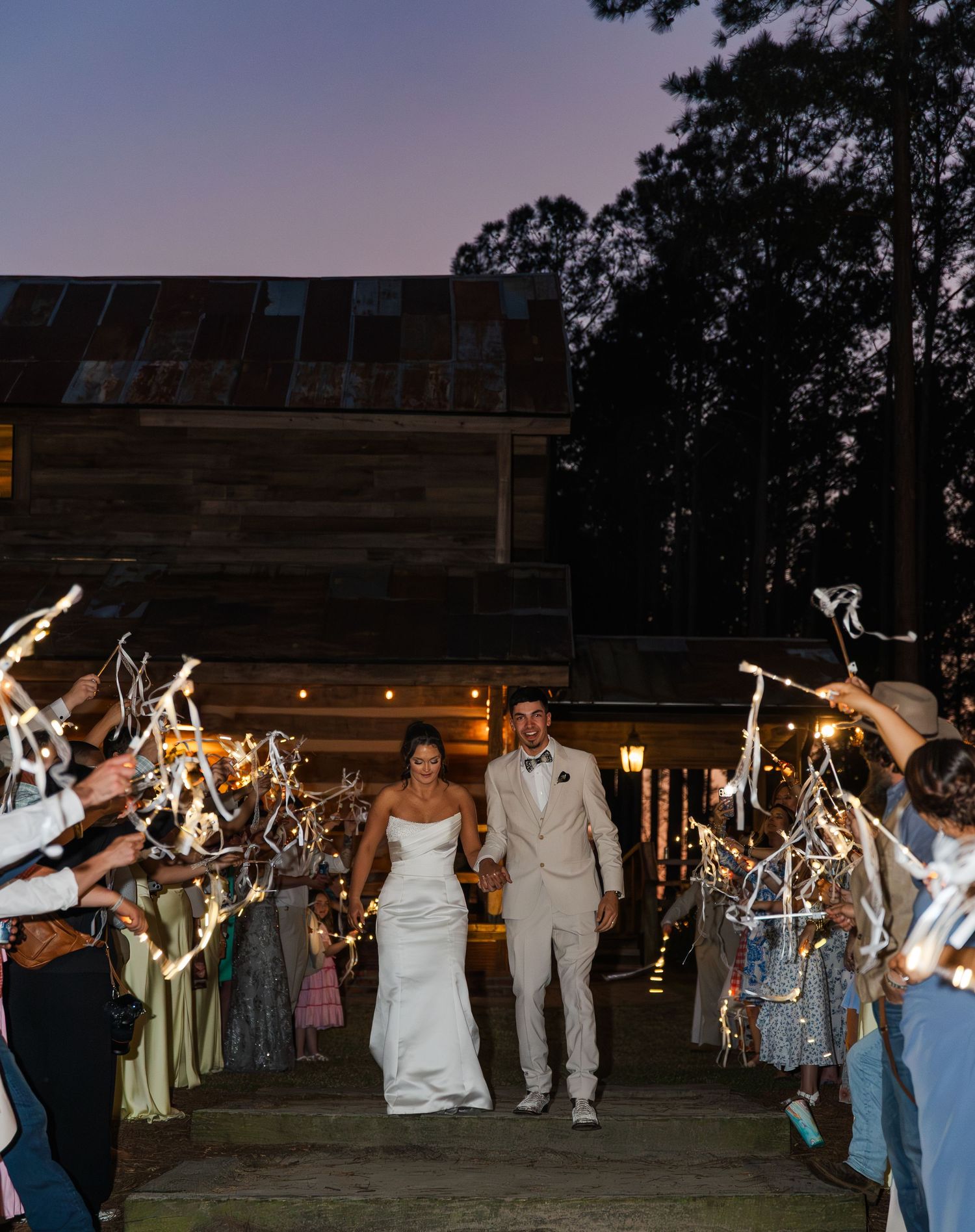 Newlyweds exit their rustic barn wedding reception through a sparkler send-off tunnel at twilight.